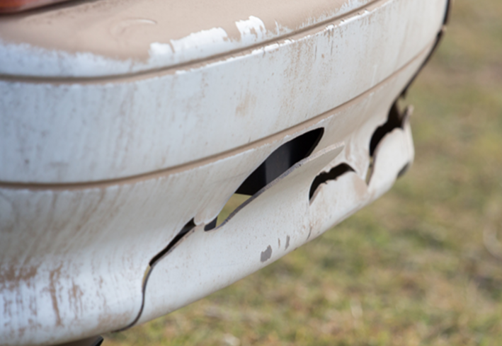 Close-up of a rusty, damaged white car bumper with cracks and peeling paint, on a grassy surface.