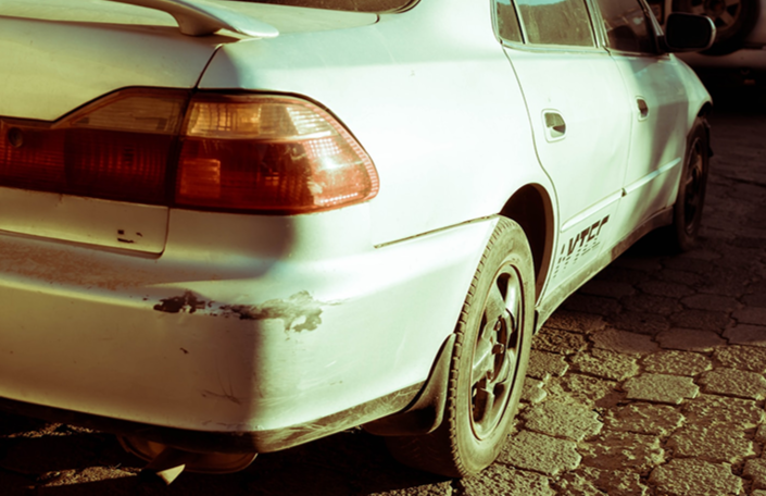 The rear side of an old white sedan with visible scratches and damage, parked on a cracked asphalt surface.