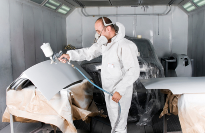 A person in protective gear spray-paints a car part in a spray booth.