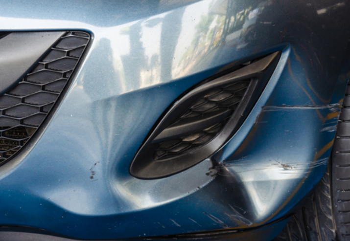 Close-up of the front left side of a blue car, showing the headlight, part of the grille, and the front bumper with some scratches and dirt.