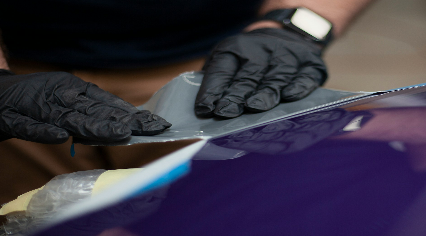 Person wearing black gloves applying a vinyl wrap to a car's hood.