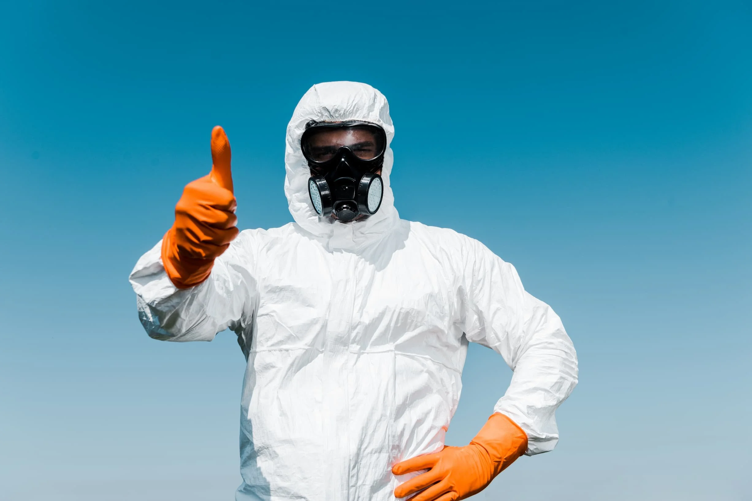 A person wearing a white hazmat suit, black gas mask, orange gloves, giving a thumbs up against a clear blue sky.