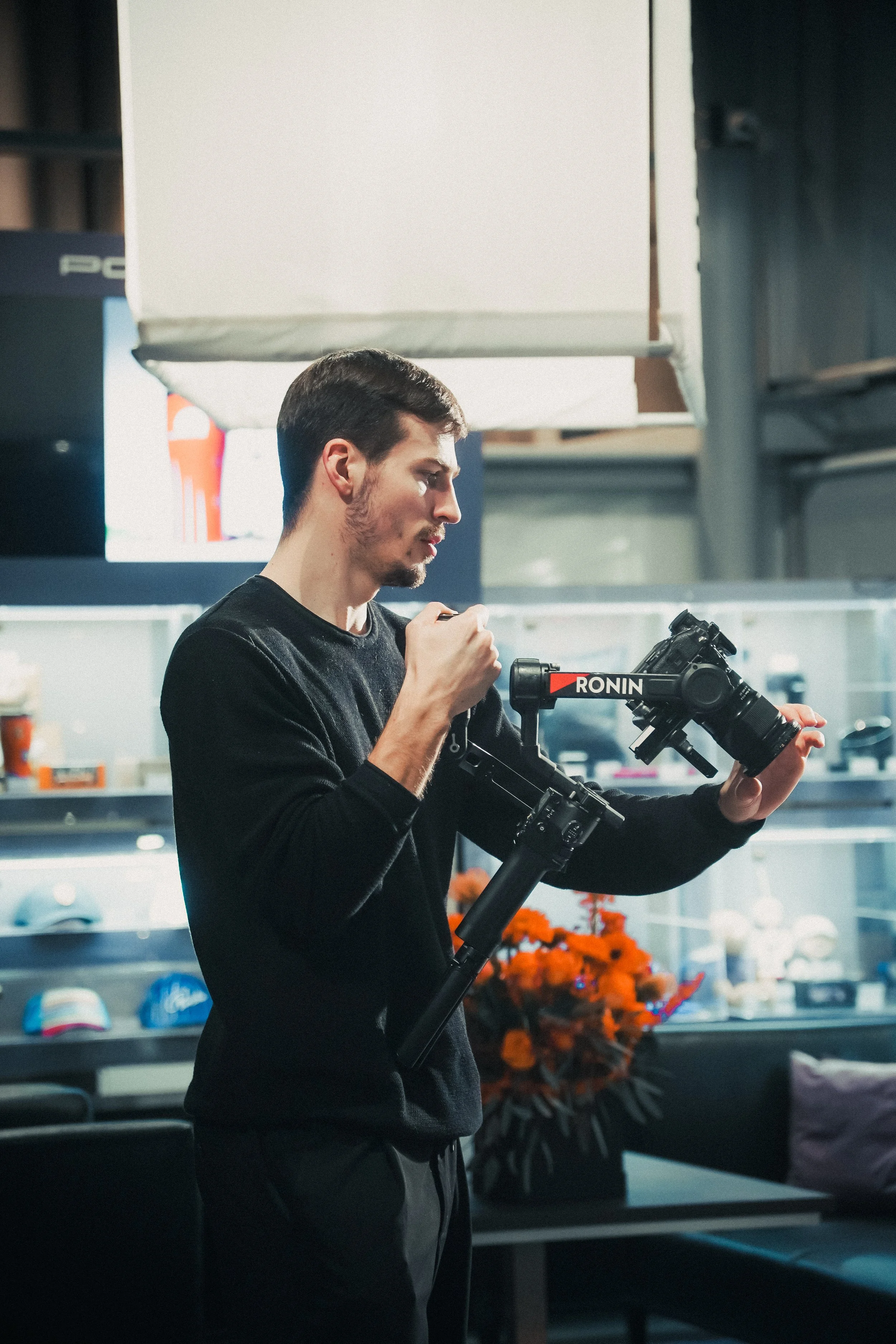 A man in a black long-sleeve shirt is adjusting a camera mounted on a stabilizer in a retail store or showroom with hats and accessories displayed on shelves behind him.