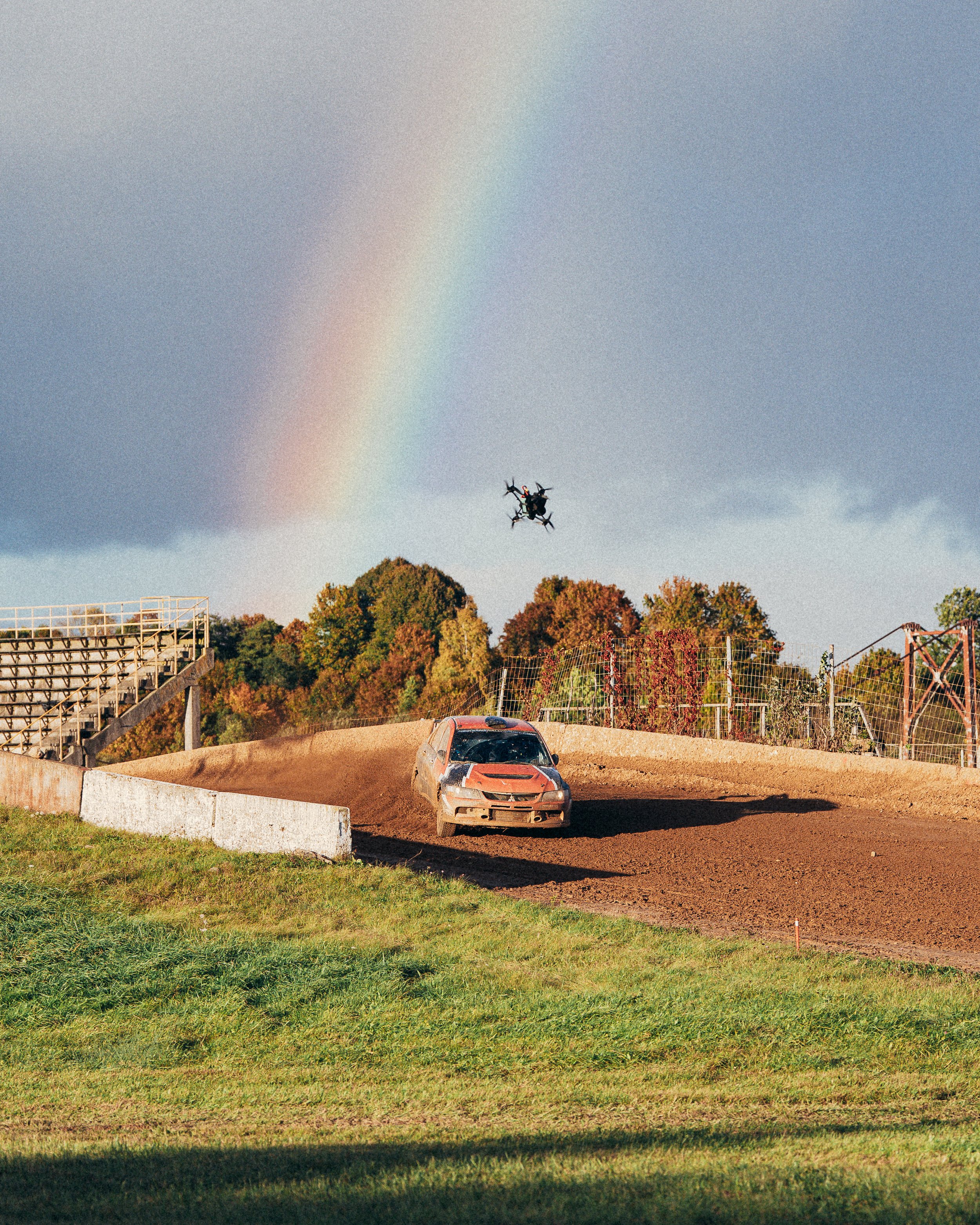 A dirt track racecar flying off a jump with a drone overhead, rainbow in sky, trees in background.