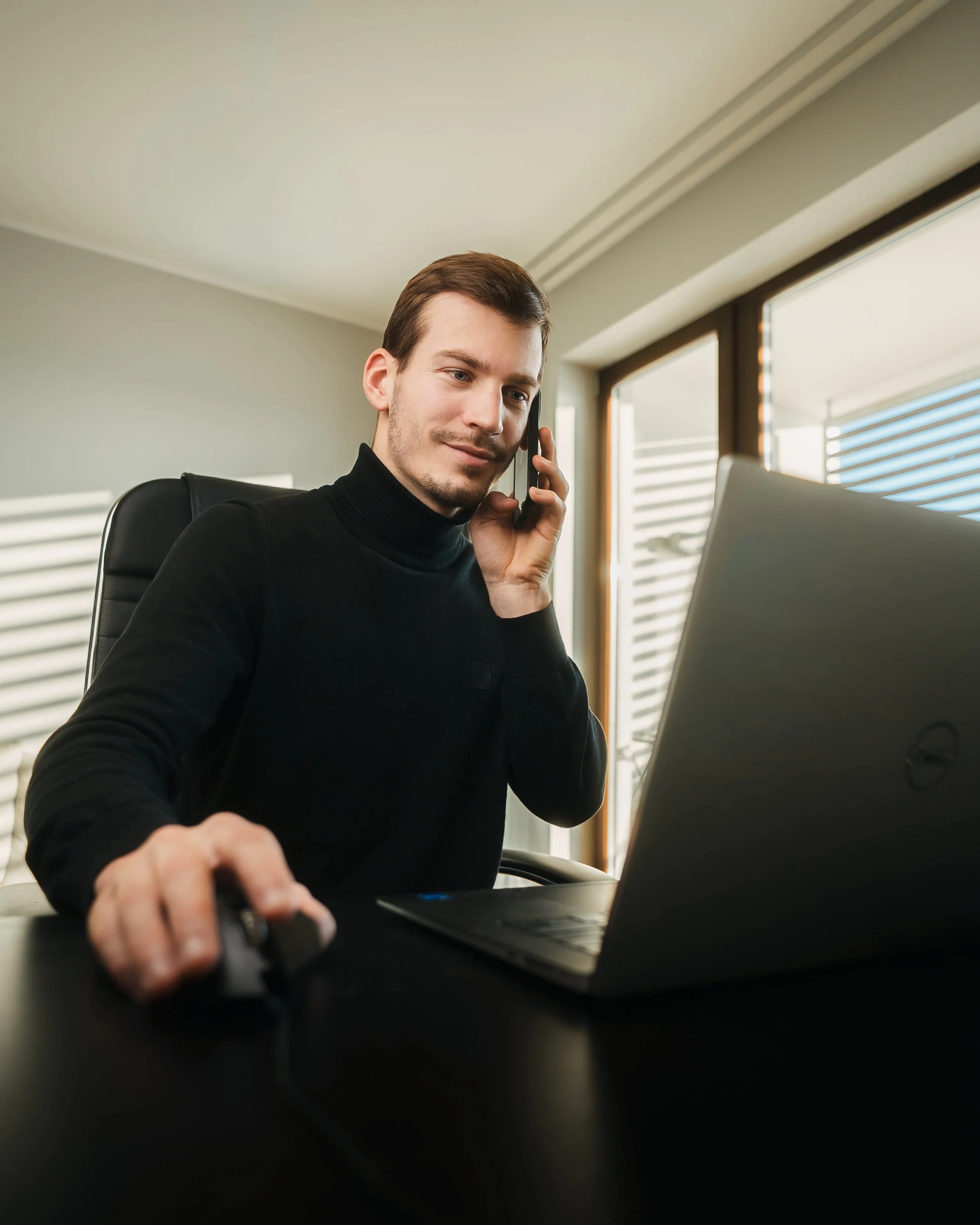 A young man sitting at a desk, using a laptop and talking on a cell phone in a modern office with large windows.
