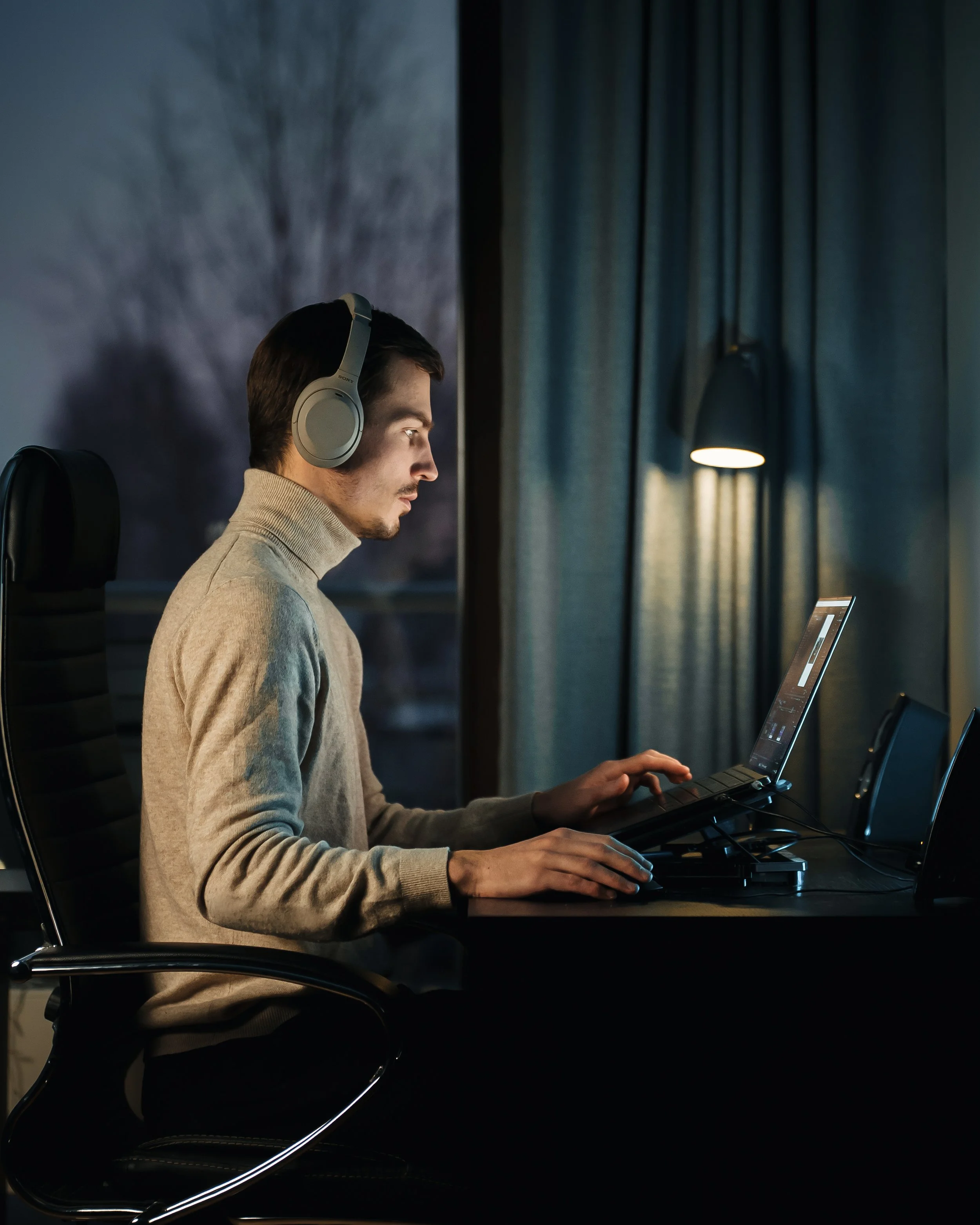 A young man wearing headphones is sitting at a desk working on a laptop in a dimly lit room with a window, curtain, and a small lamp.