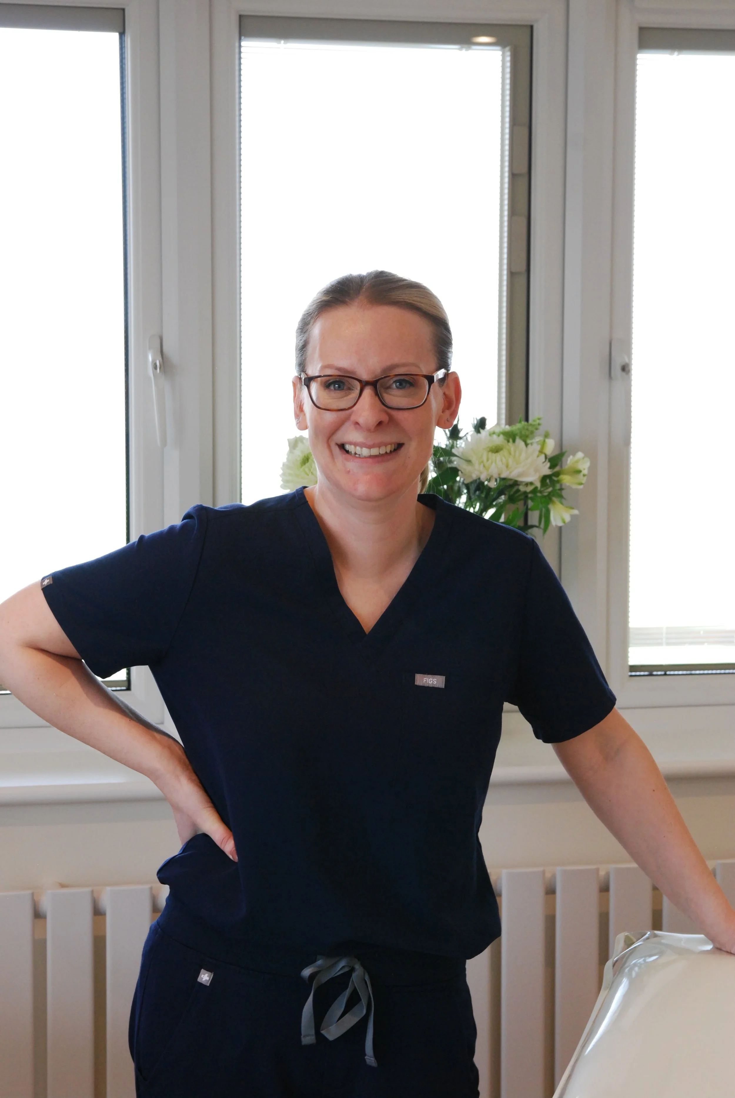 A smiling woman in navy scrubs with glasses standing indoors near a window with a white flower arrangement in the background.