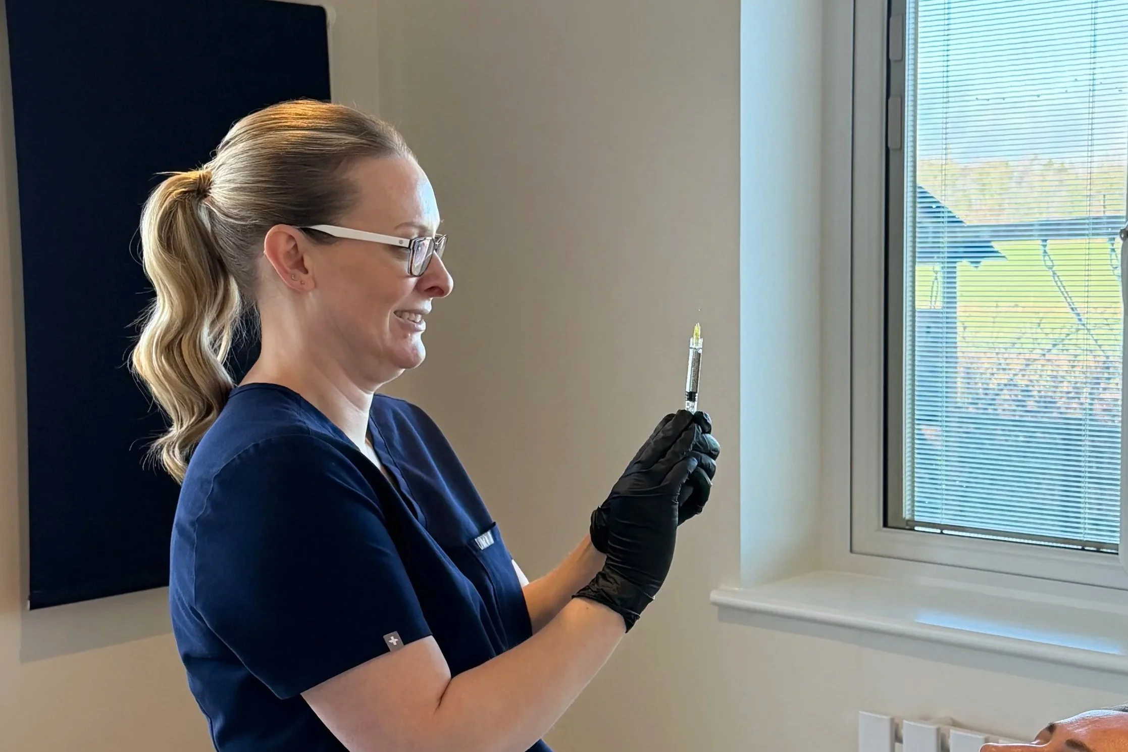 Nurse in navy scrubs and black gloves holding a syringe, smiling while preparing a vaccination, standing near a window with outside sunlight.