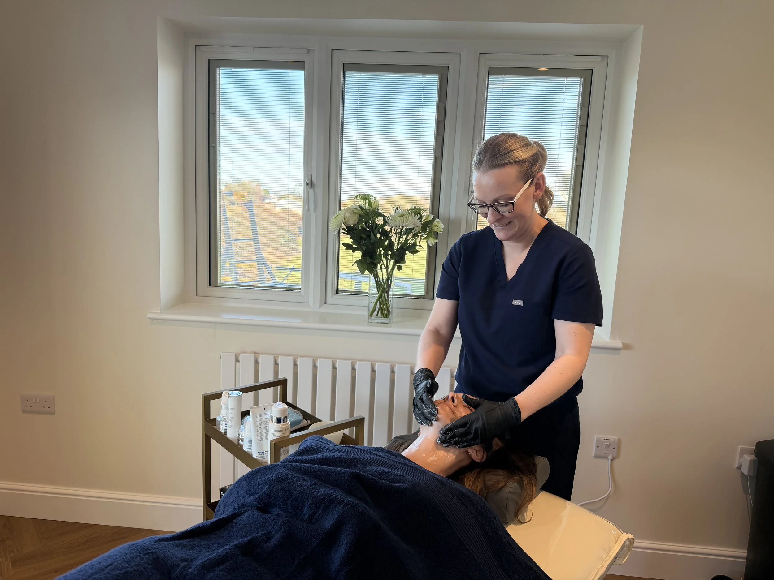 A woman in black scrubs and glasses is performing a facial treatment on a person lying on a treatment bed in a well-lit room with a window and a vase of flowers.