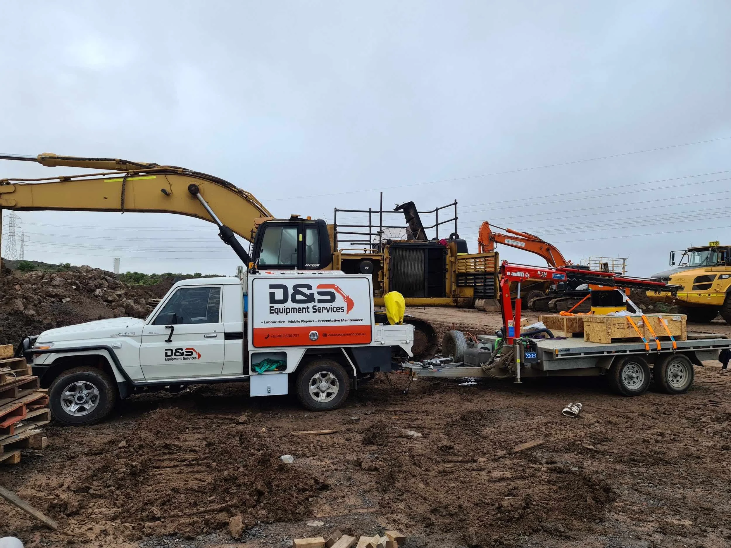 Construction site with a white D&S Equipment Services truck, excavators, and construction materials on muddy ground under a cloudy sky.