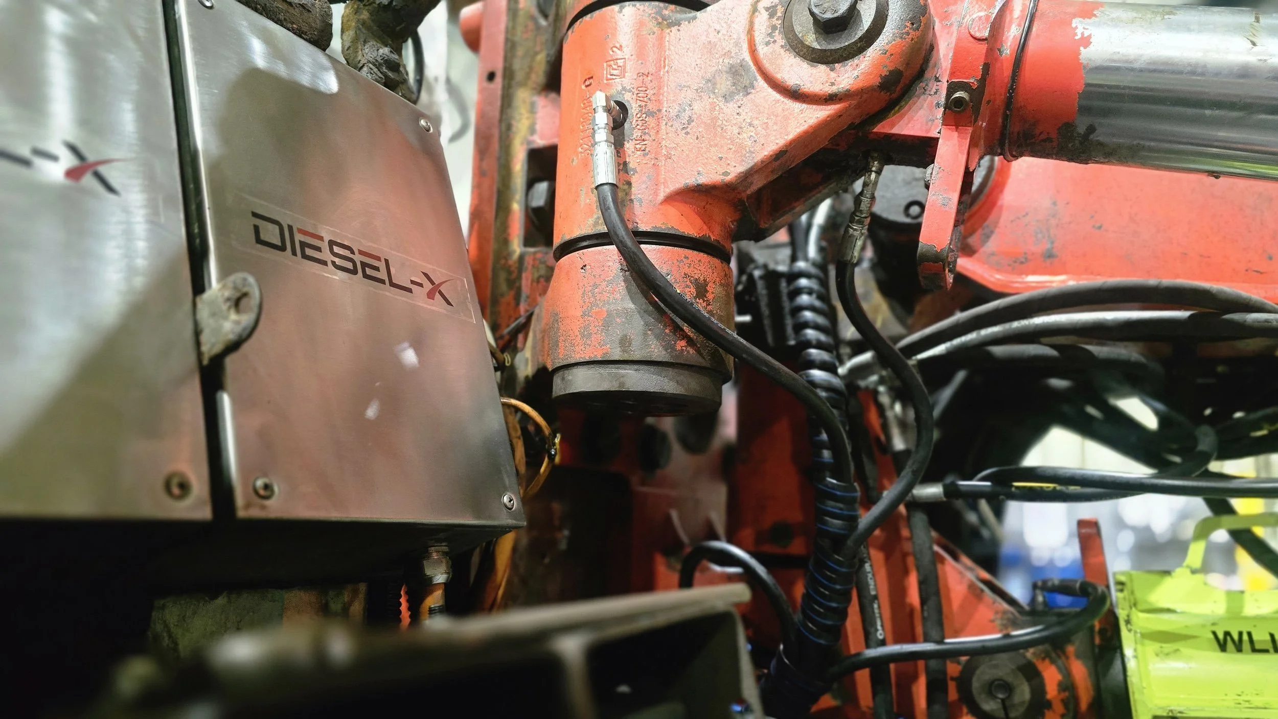 Close-up of a mechanical engine component with a DIESEL-X branded metal cover, connected hoses, and metal parts with visible bolts and wiring.