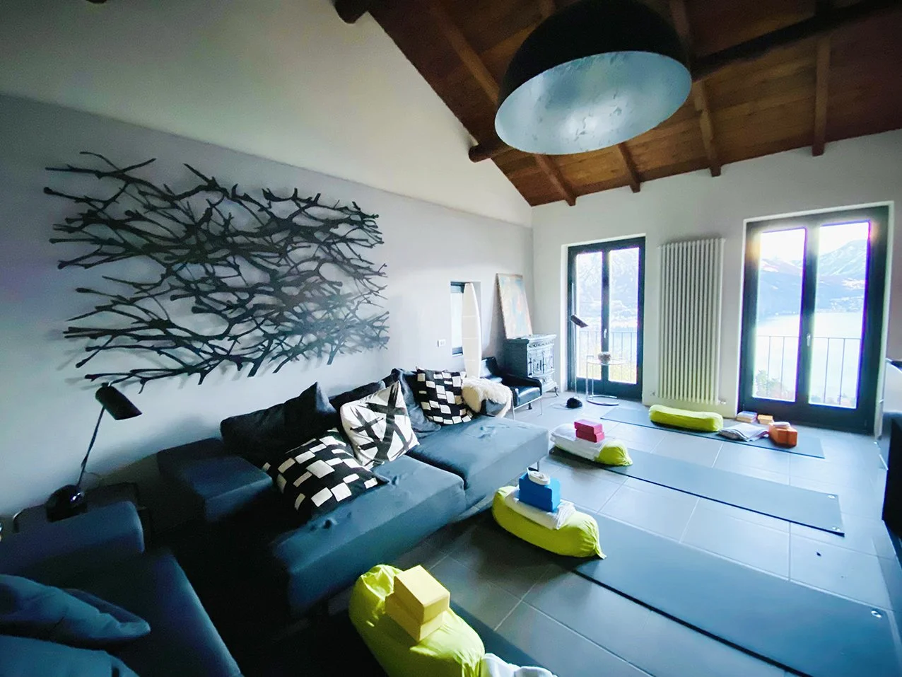 Wohnzimmer mit couch, Teppich, Yoga-Matten, Wasserflaschen, Kissen, einer großen Fensterfront mit Blick auf Berge und See, Holzdecke, moderner Beleuchtung und Kunstwerk an der Wand.