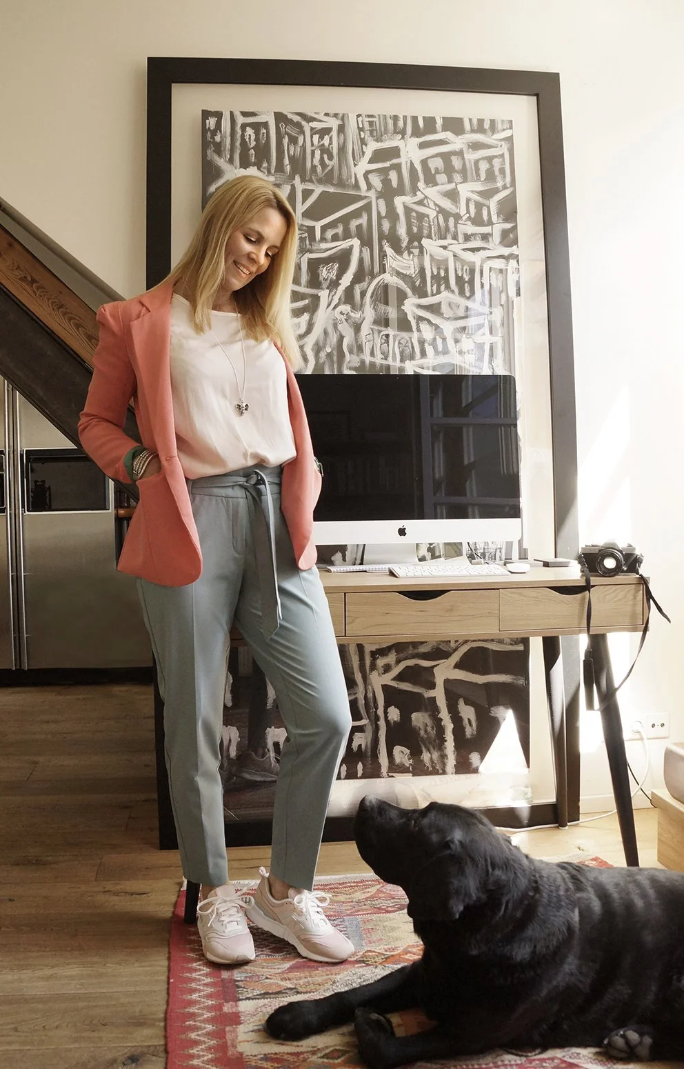 Eine lächelnde Frau steht in einem Wohnzimmer, trägt eine rosa Blazer, eine weiße Bluse, graue Hosen mit Gürtel und weiße Turnschuhe. Vor ihr liegt ein schwarzer Hund, der auf dem Boden liegt. Im Hintergrund ein Schreibtisch mit einem Computer, eine Kamera und ein schwarzes und weißes Kunstwerk.