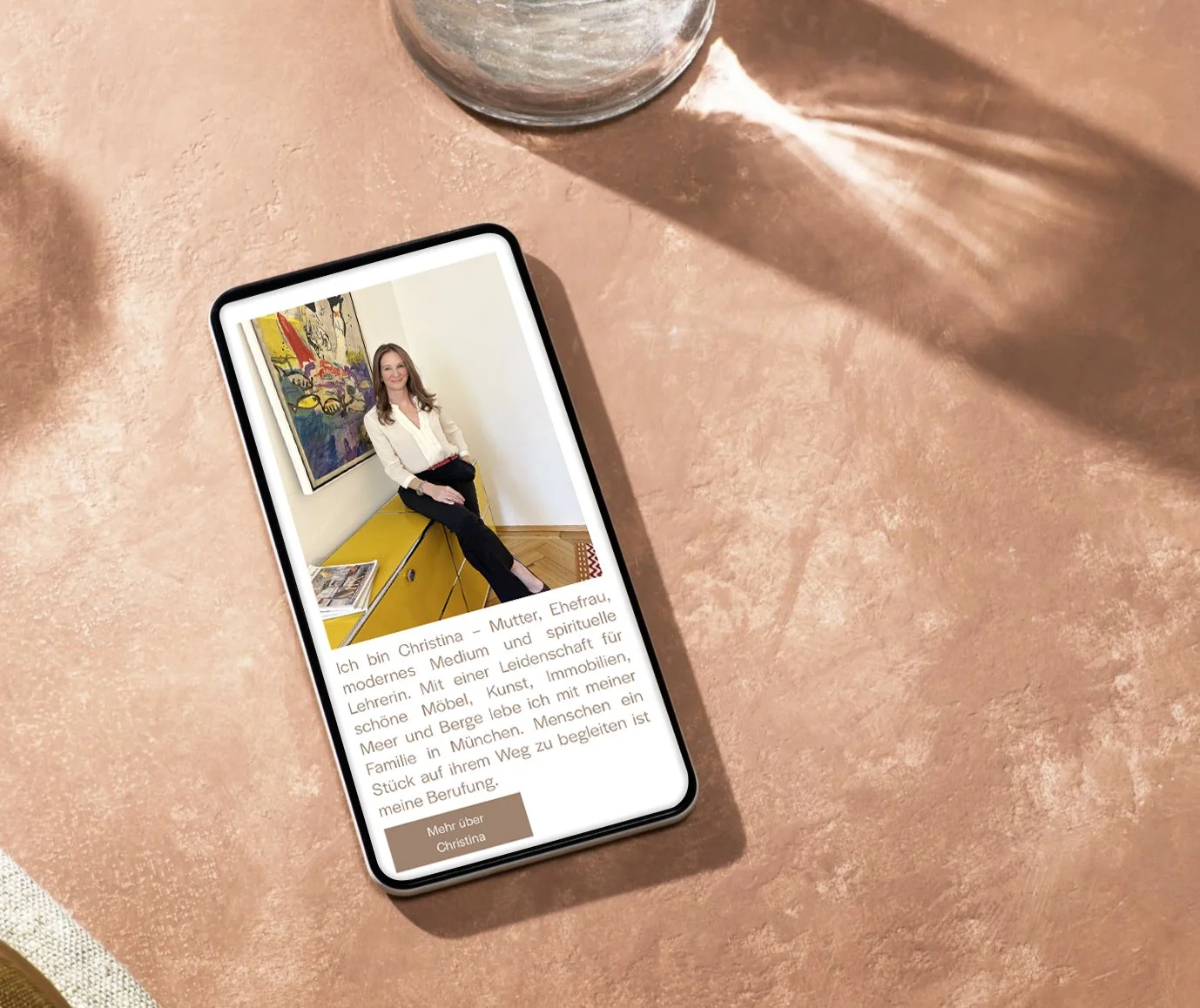 Smartphone auf einem Tisch zeigt eine Frau namens Christina, sitzend vor Kunstwerken, mit einem Text über sie auf Deutsch.