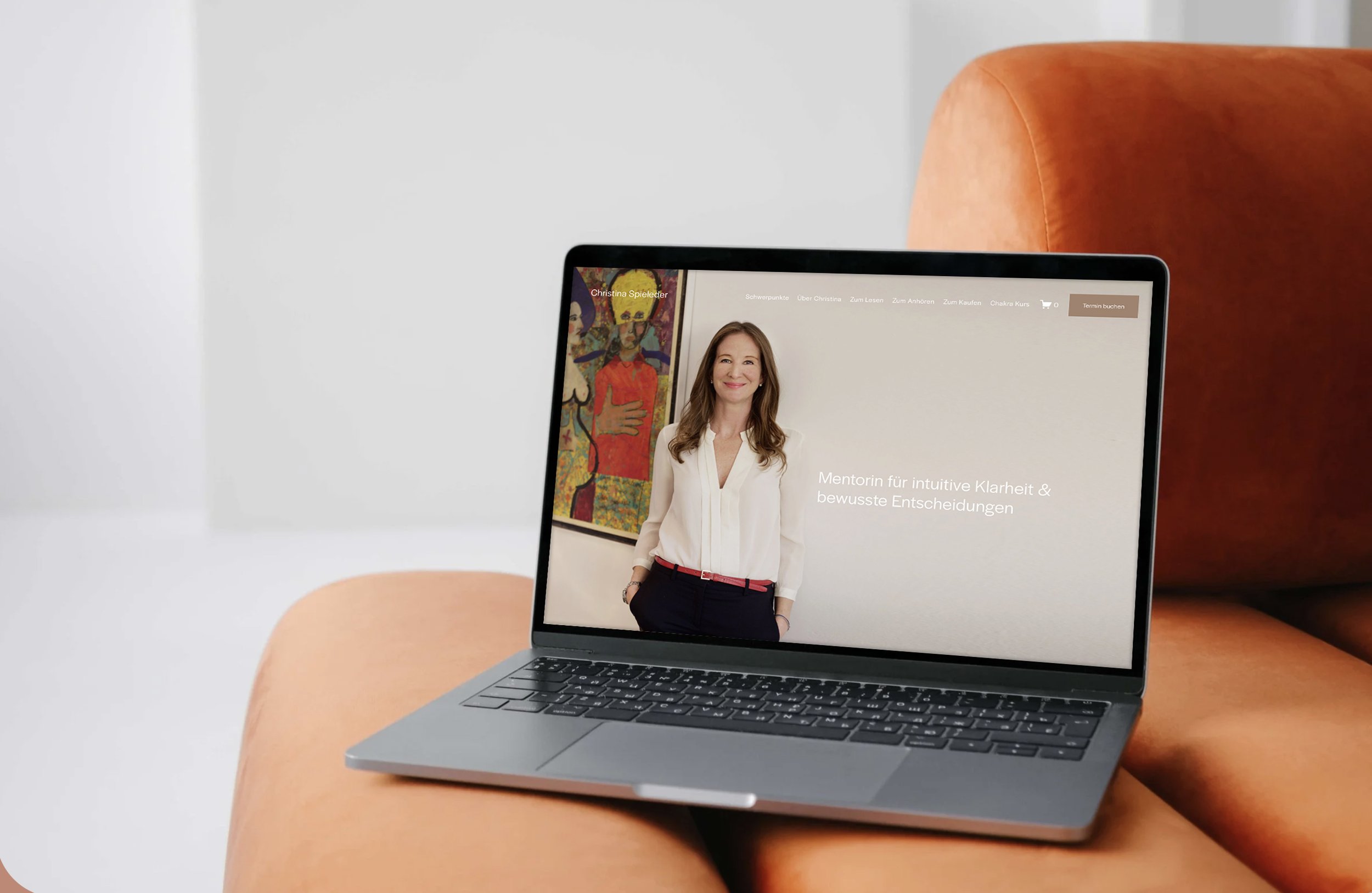 Laptop auf einem orangefarbenen Sofa mit Webseite einer Mentorin für Klarheit und bewusste Entscheidungen, neben einem Gemälde an der Wand.