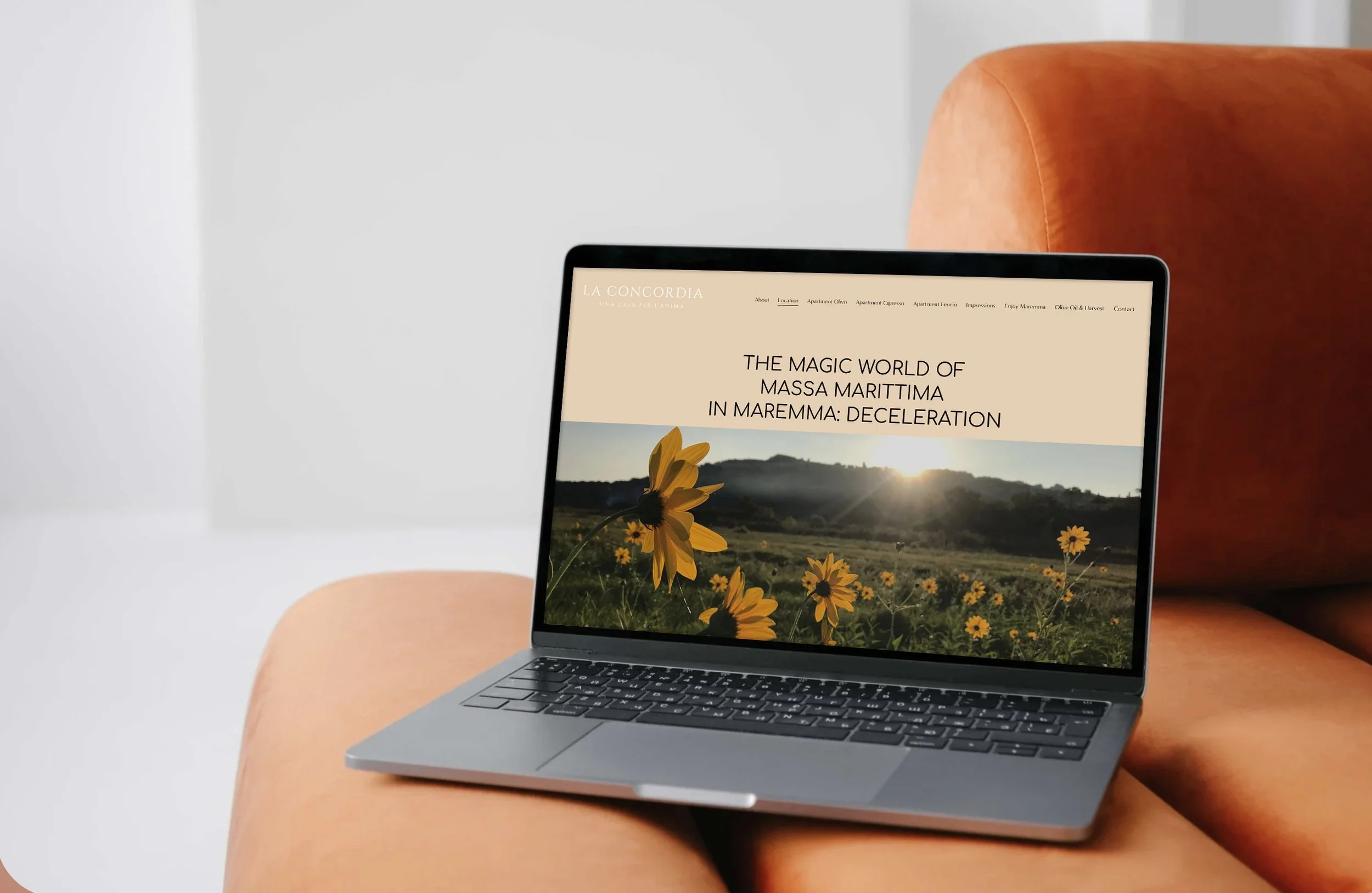Ein Laptop liegt auf einem orangefarbenen Stuhl. Auf dem Bildschirm ist eine Webseite mit einem Bild von Sonnenblumen vor einem Sonnenuntergang und dem Text "The magic world of Massa Marittima in Maremma: Deceleration" zu sehen.