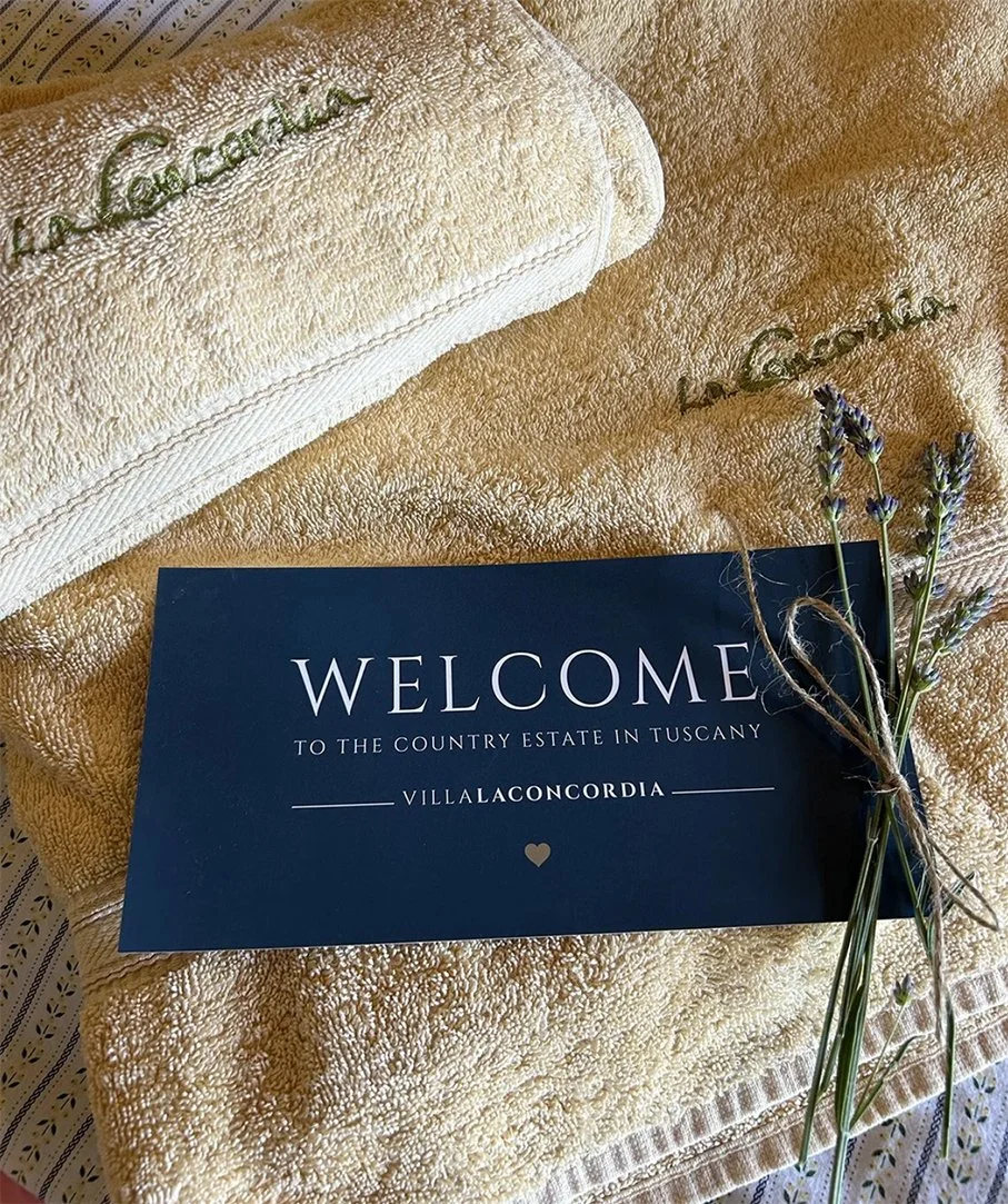 Ein Willkommensschild mit der Aufschrift 'Welcome to the country estate in Tuscany Villla Concordia', gelegen auf einem gelben Handtuch, dazu Lavendel und eine Handtuchrolle.