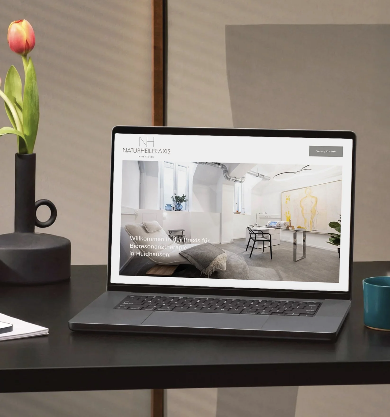 Ein offener Laptop auf einem schwarzen Tisch zeigt eine Website für eine Naturheilpraxis in Haidhausen, mit einem Behandlungsraum im Hintergrund. Auf dem Tisch stehen eine schwarze Vase mit Tulpen, ein Notizbuch, und eine blaue Tasse.