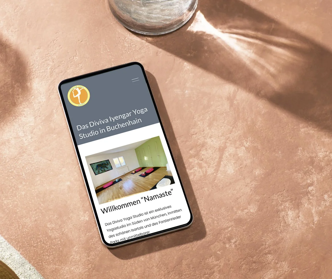 Smartphone liegt auf einem rosa Tisch, zeigt eine Webseite für das Diviva Iyengar Yoga Studio in Buchenhain, mit einem Bild des Yogastudios und einem Begrüßungstext.
