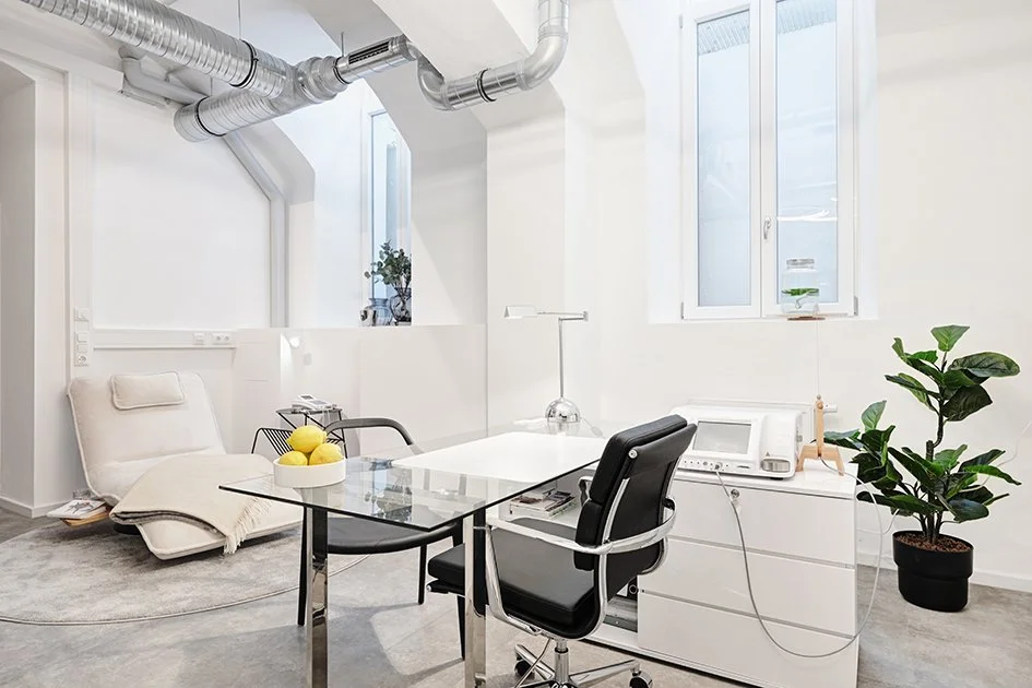 Ein modernes, helles Büro mit weißer Wand, großen Fenstern, einem Schreibtisch, zwei Bürostühlen, einer Pflanzen, einem Schreibtisch mit Computer und anderen Büroartikeln, sowie einem Relaxsessel mit Decke.