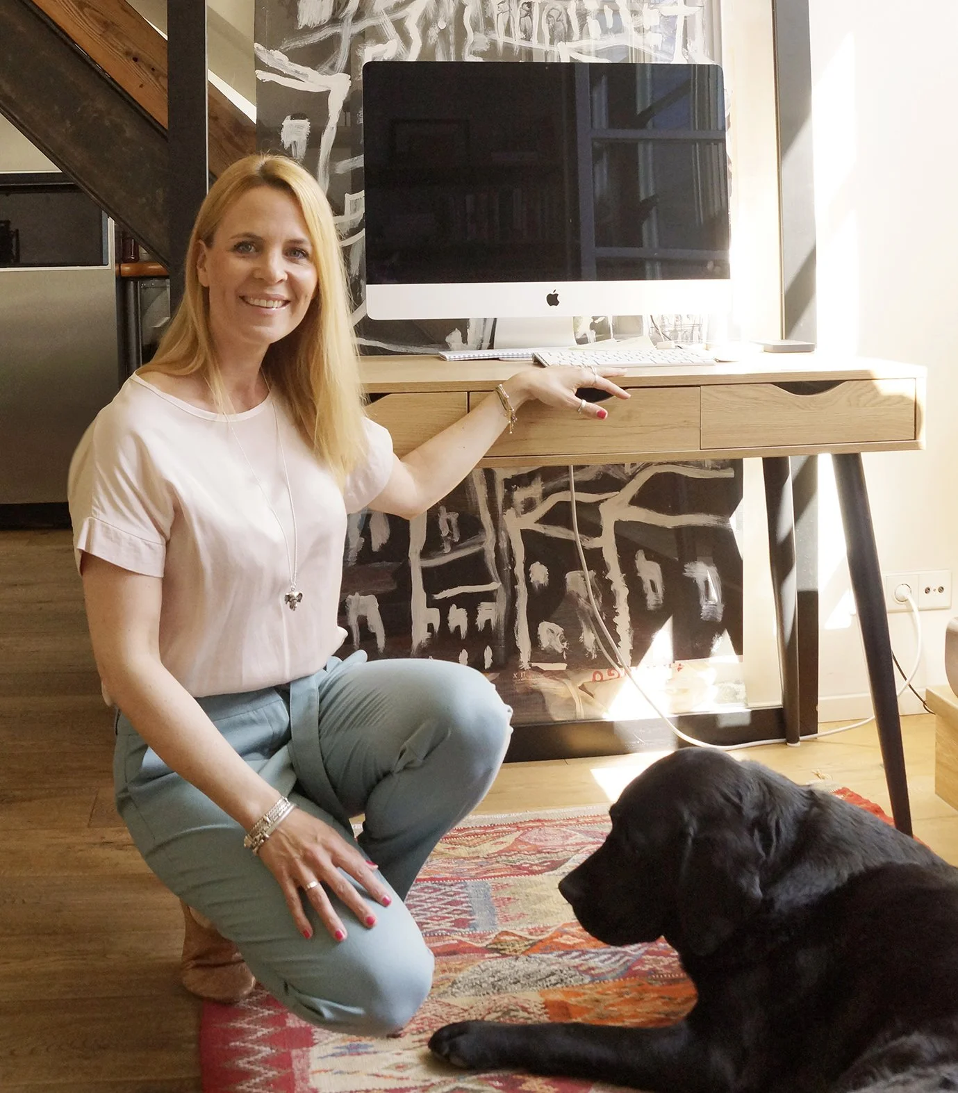 Eine Frau mit langen blondem Haar kniet neben einem schwarzen Hund auf einem roten Teppich in einem modernen Raum. Im Hintergrund steht ein weißer Computer auf einem Holztisch, neben einem dunklen Wandbild.