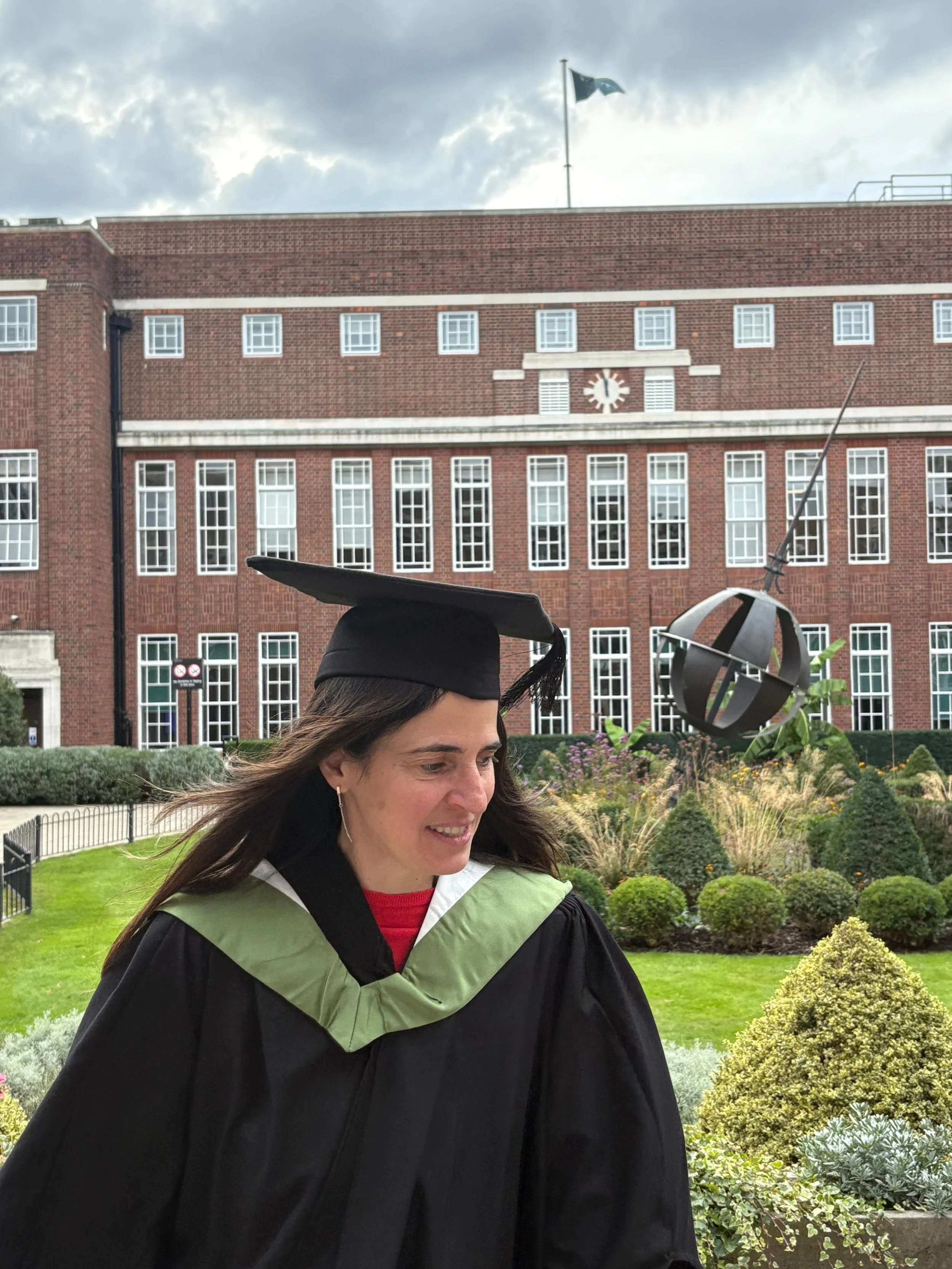 Vrouw in afstudeerkleding met academisch hoedje, buiten voor een roodstenen gebouw met ramen en een tuin, tijdens een grijze zonnige dag.
