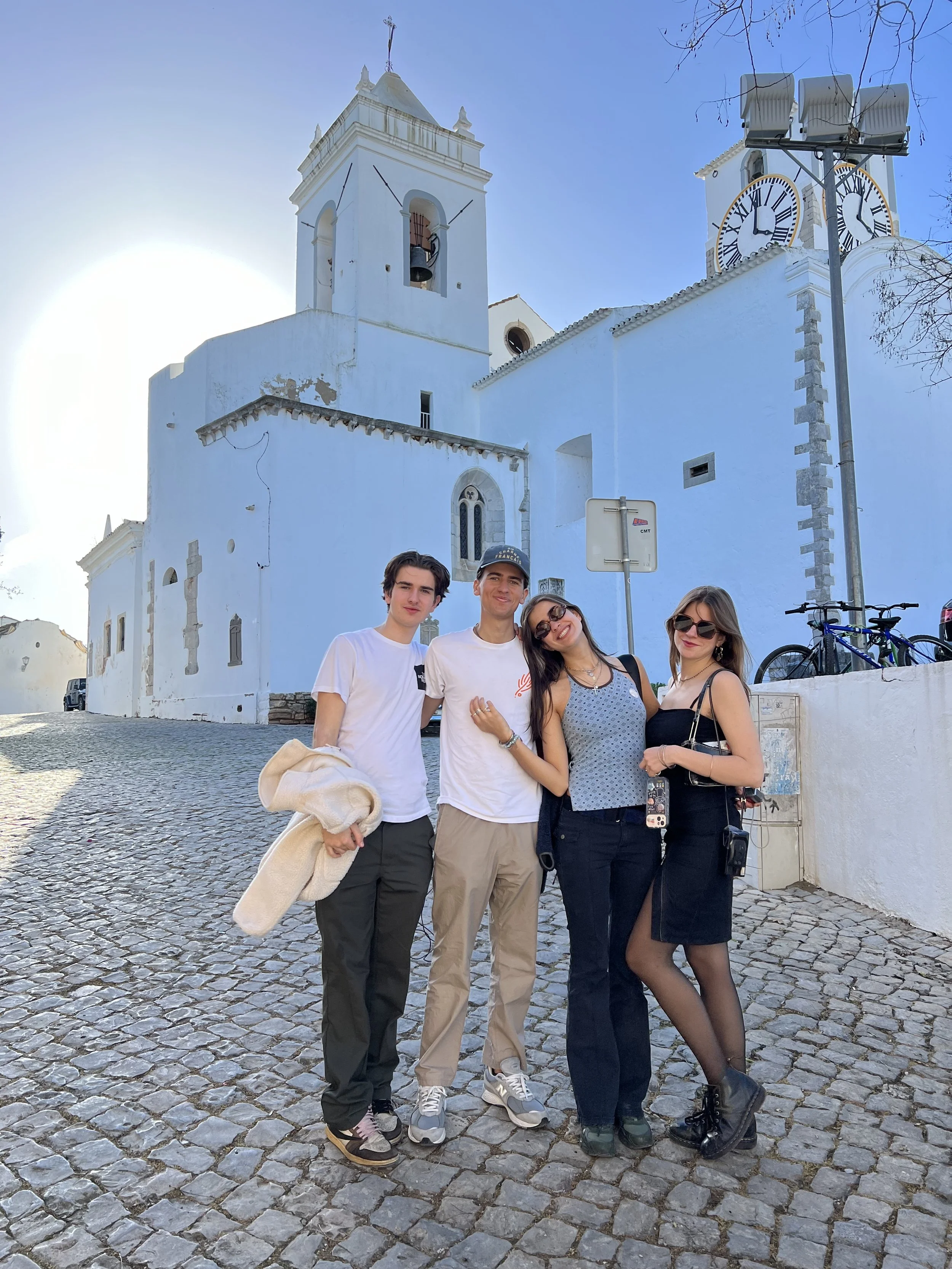 Vijf mensen poseerden op een stenen plein voor een witte kerk met een klok en twee urenwerken, onder een heldere blauwe hemel.