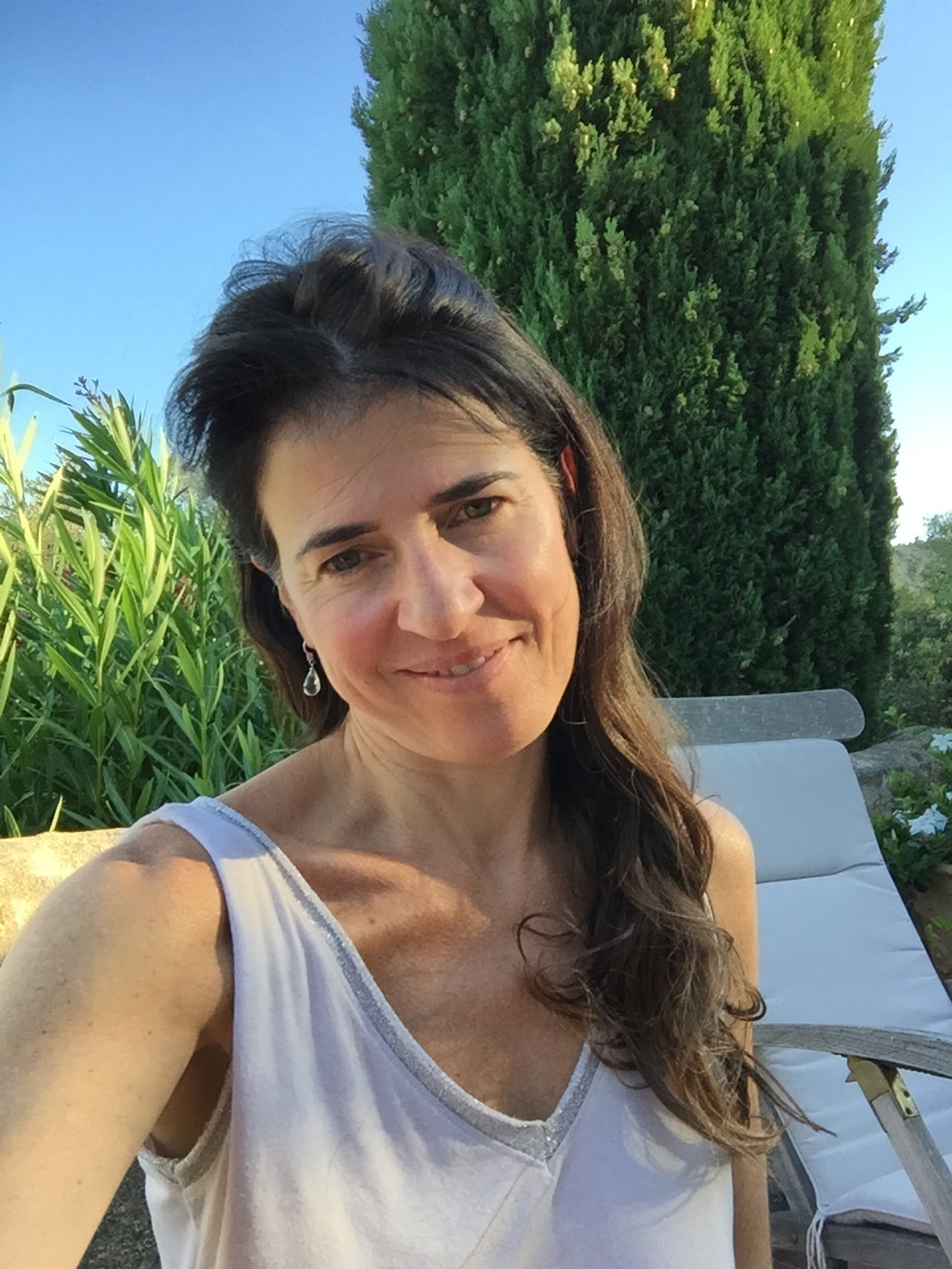 Vrouw die buiten in de zon poseert, met groene planten en een hoge boom op de achtergrond.