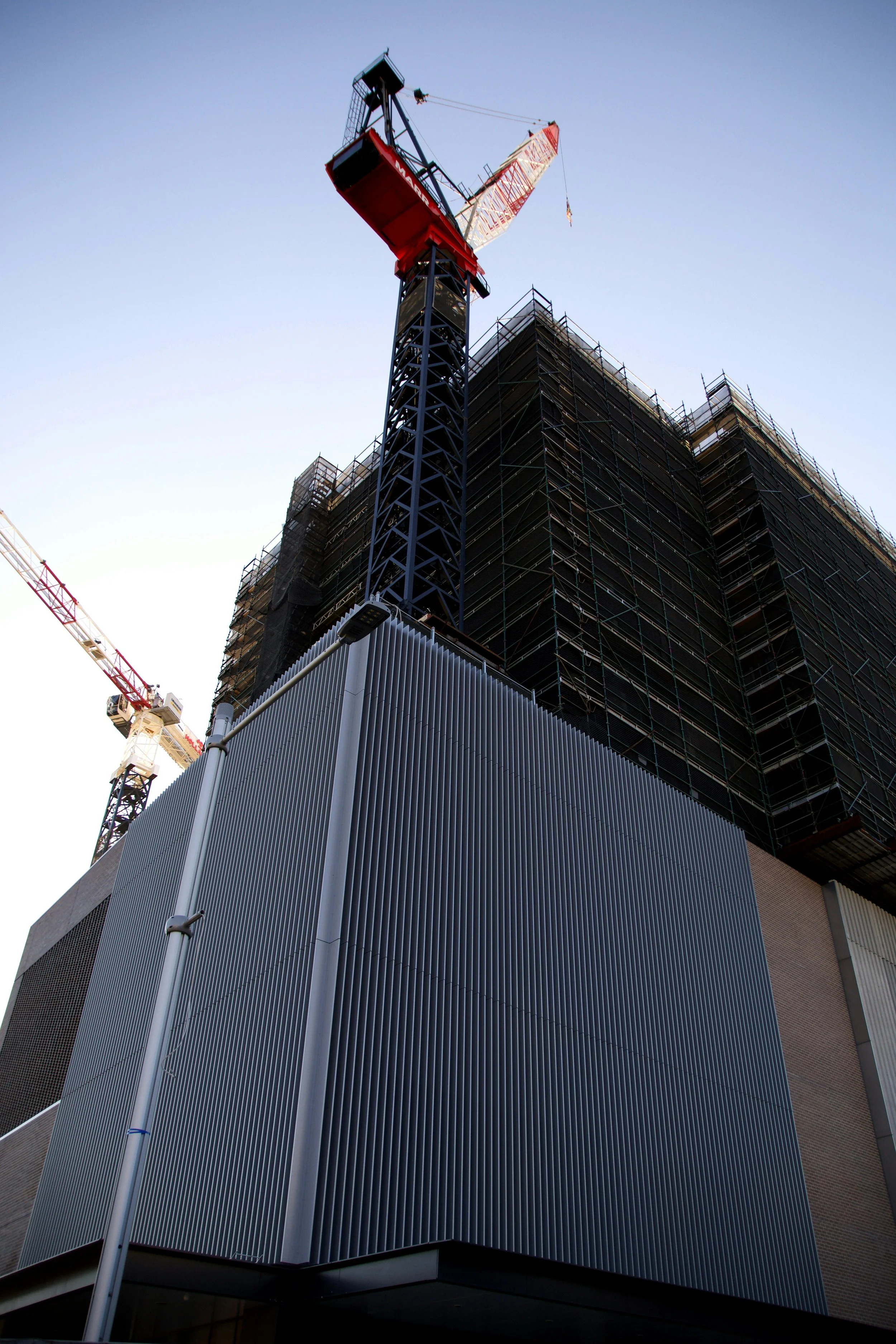A construction site with a tall crane and a building under construction, surrounded by scaffolding and partially covered with dark protective netting.
