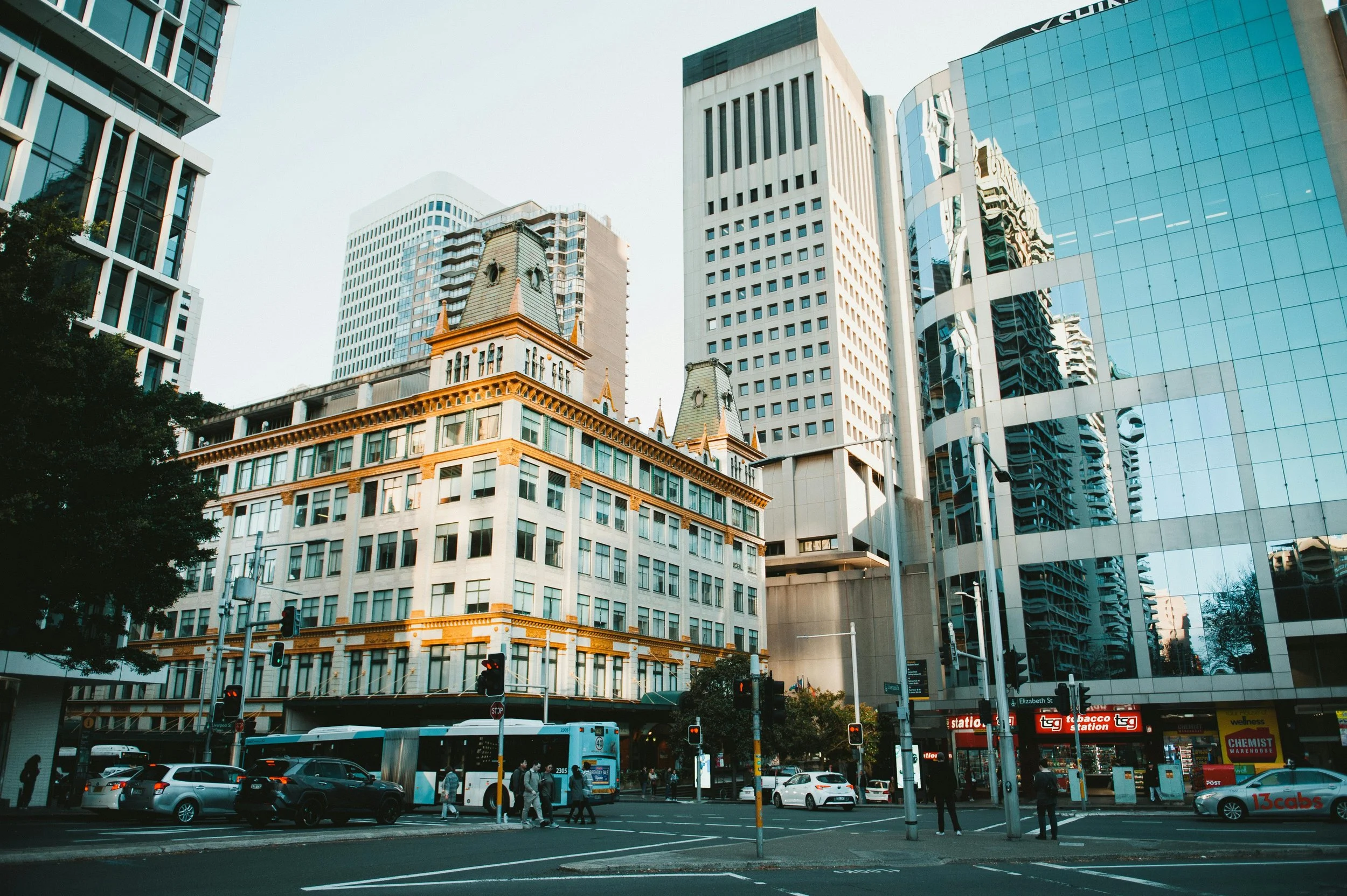 City street scene with tall modern glass buildings and an old-style building with towers. People walk, cars and a bus are on the street, and traffic lights are visible.