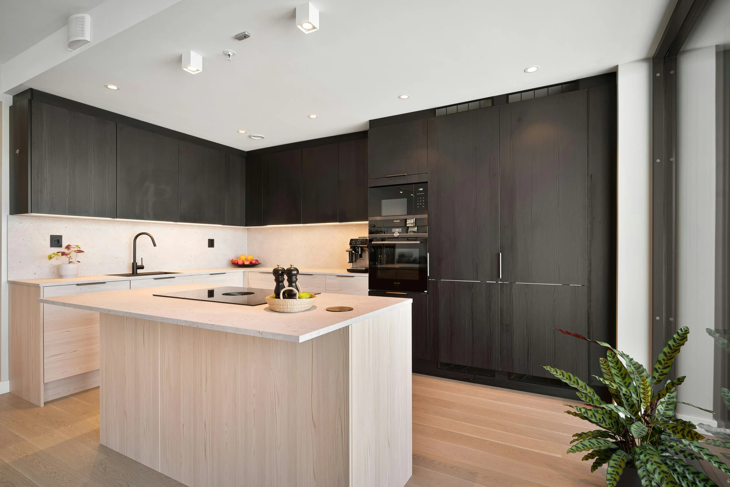 Modern kitchen with light-colored island, dark upper and tall cabinets, built-in oven, coffee machine, and minimal decor, with light wood flooring and natural light.