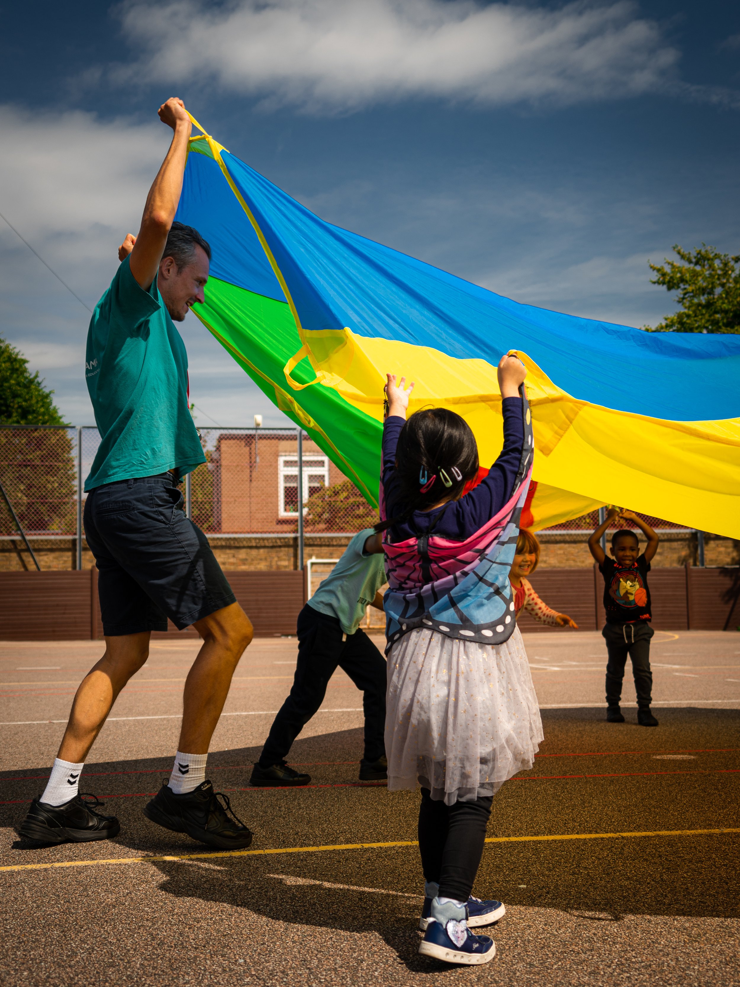 Activity Leaders and children from Global Camps playing with a colourful parachute on a school playground.