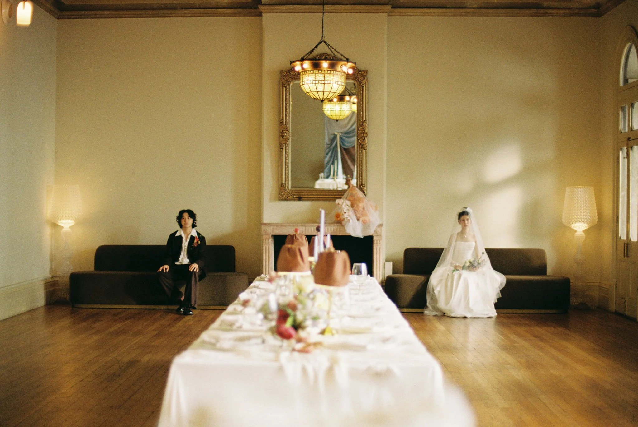 A wedding reception setup in a spacious room with two women sitting on opposite sides of a fireplace, one in a black suit and the other in a wedding dress. The table is decorated with flowers and desserts. The room is lit with warm lighting from lamps and a chandelier, and has high ceilings, large windows, and a mirror above the fireplace.