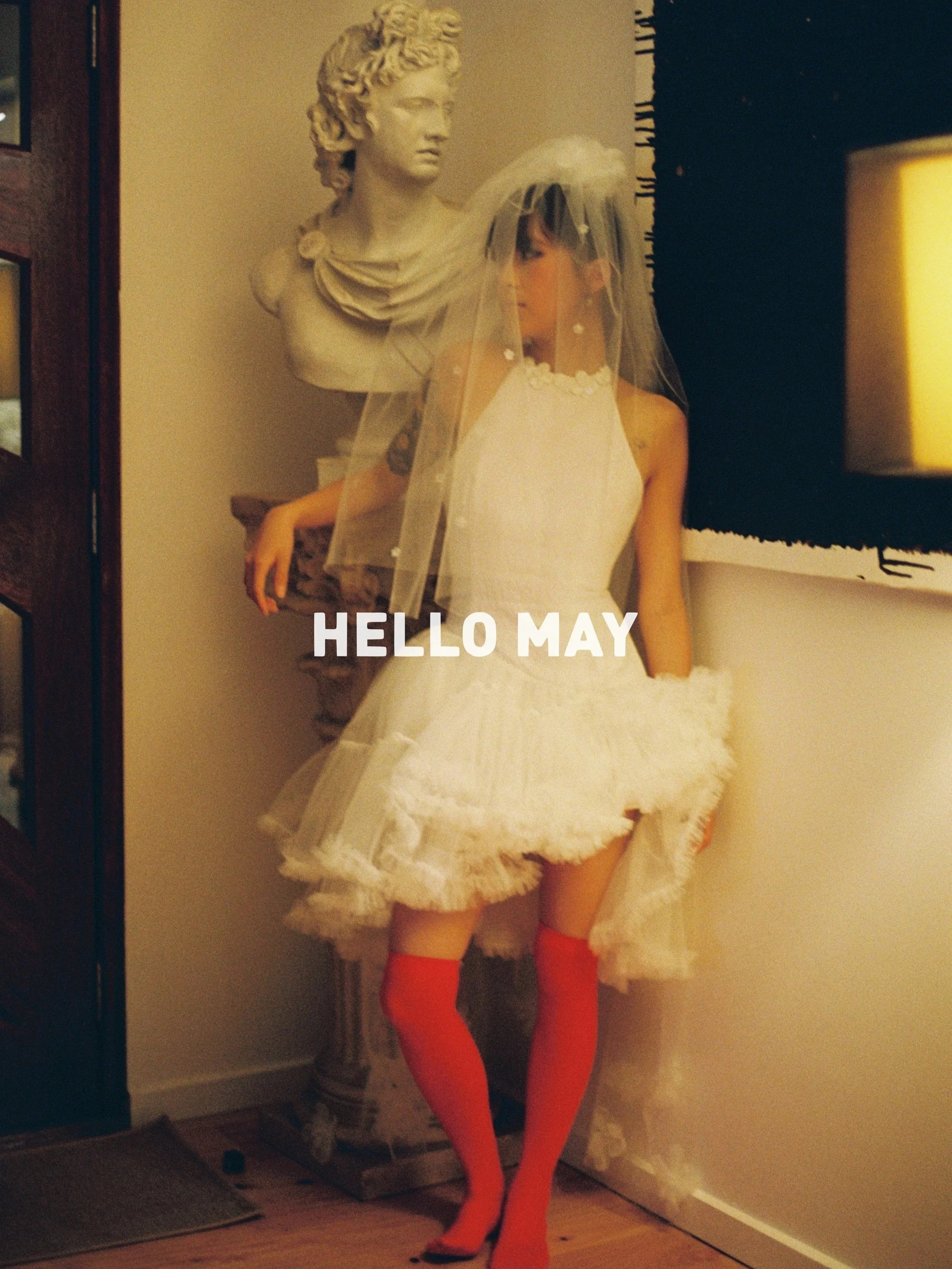 A woman dressed in a white wedding dress, orange stockings, and black high heels stands inside near a wall with her face partially visible. She wears a wedding veil. Behind her is a bust sculpture of a woman with curly hair, placed on a pedestal. The setting appears to be an indoor room with a wooden floor, a dark wall, and a paper bag or decorative piece in the background.