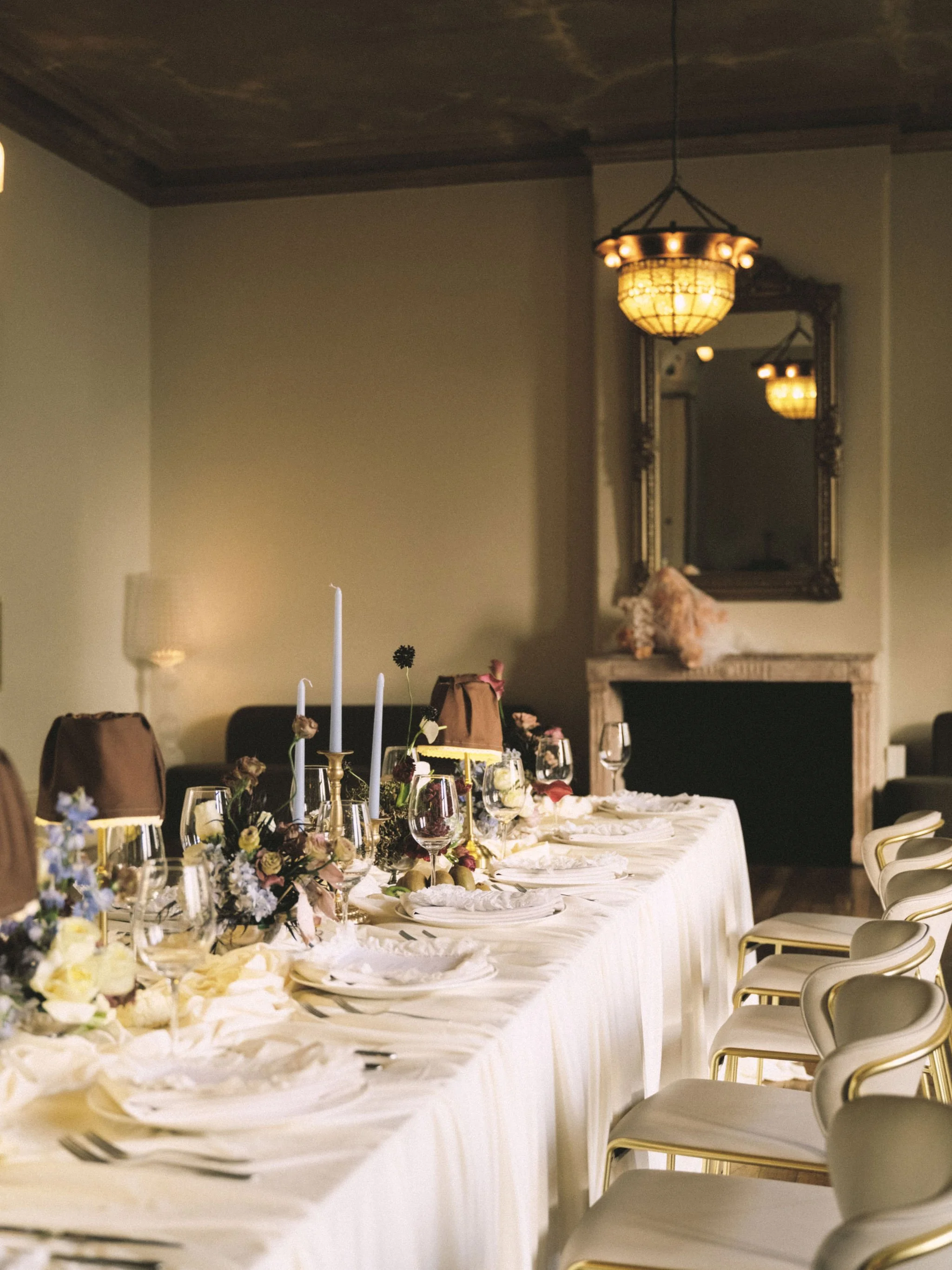 A long banquet table set for a formal event, decorated with floral arrangements, tall candles, and elegant tableware, in a room with ornate chandeliers and a fireplace with a mirror above it.