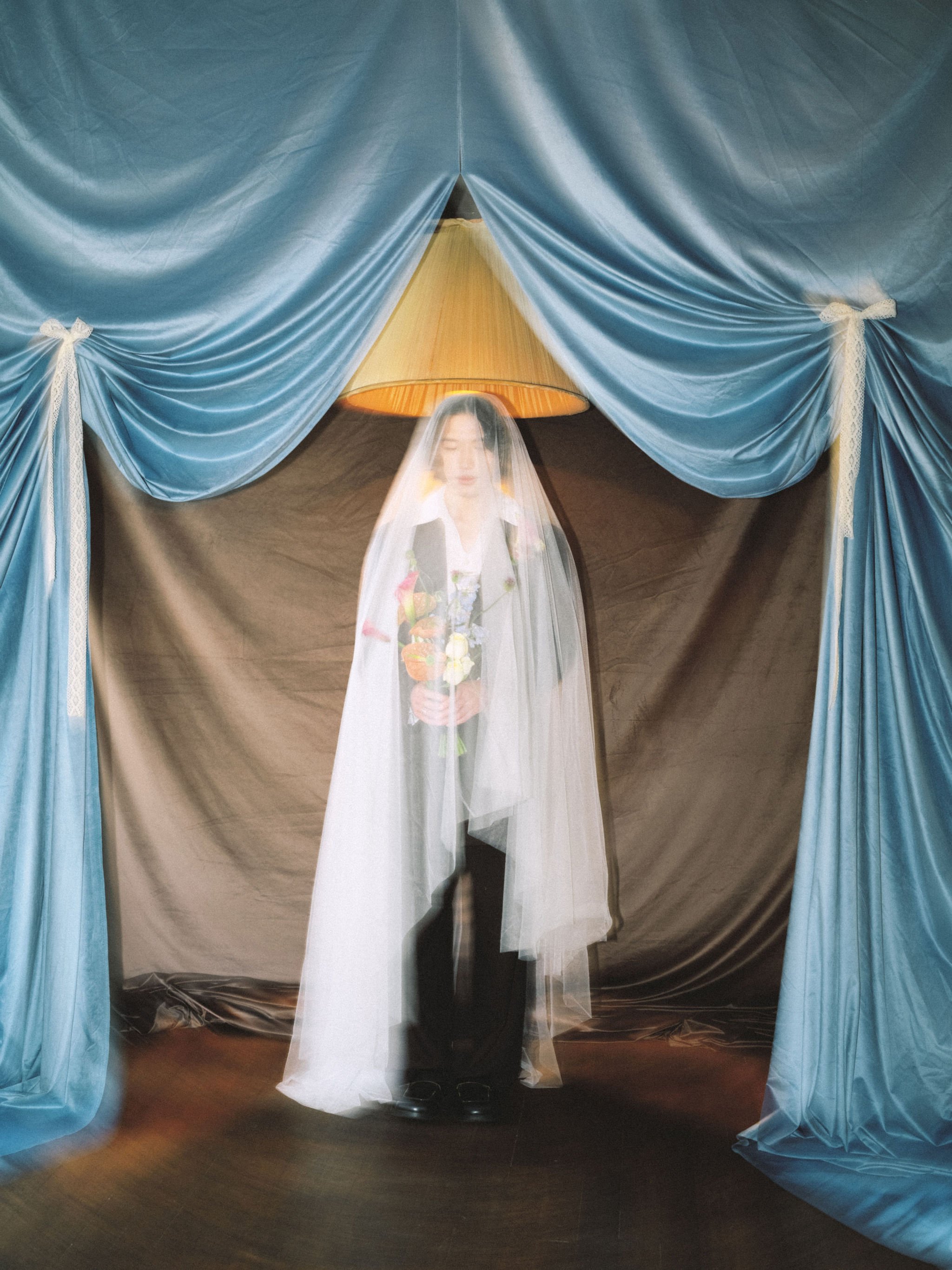 A person with a blurred face standing behind a sheer white curtain, flanked by blue drapes tied with lace bows, and a warm-colored lampshade visible above.