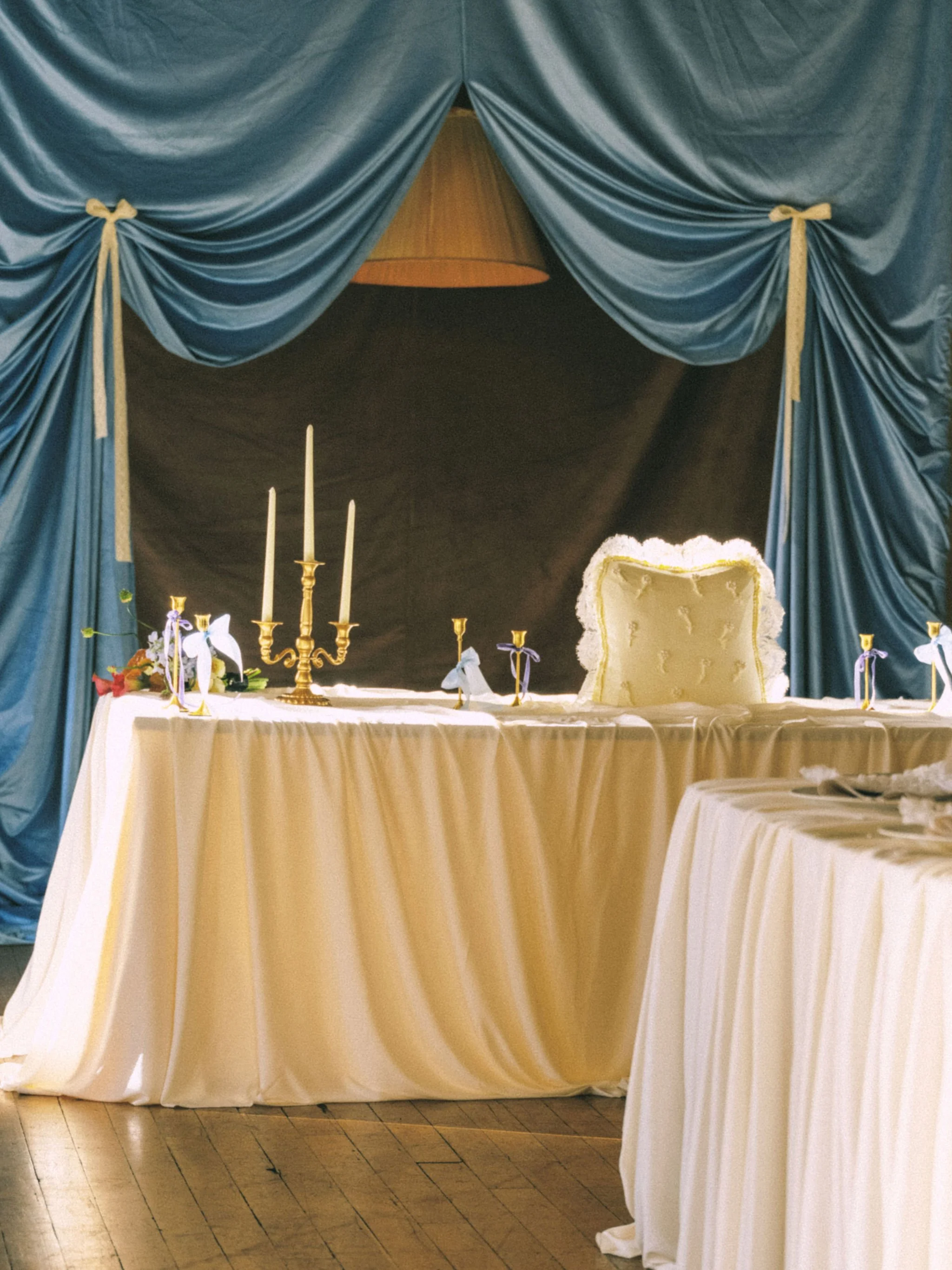 A banquet table with a white tablecloth, gold candelabra with white candles, and decorative items, set against a backdrop of dark blue curtains with gold tiebacks and an elegant chair with gold accents.