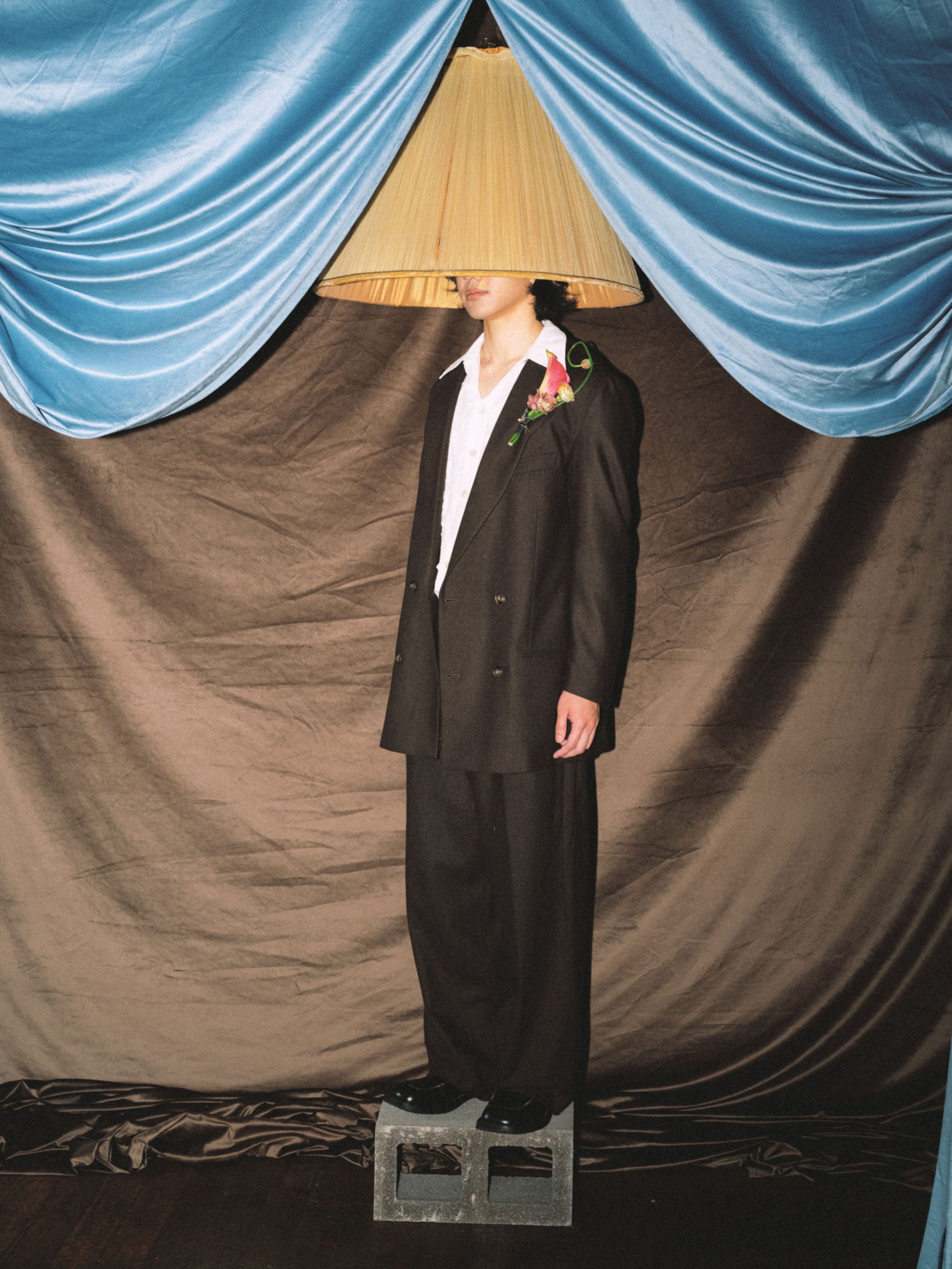 A person with black hair wearing a black blazer, white shirt, and black pants is standing on a concrete block. Their head is hidden behind a beige lampshade, creating an optical illusion. Blue velvet curtains are draped in the background.