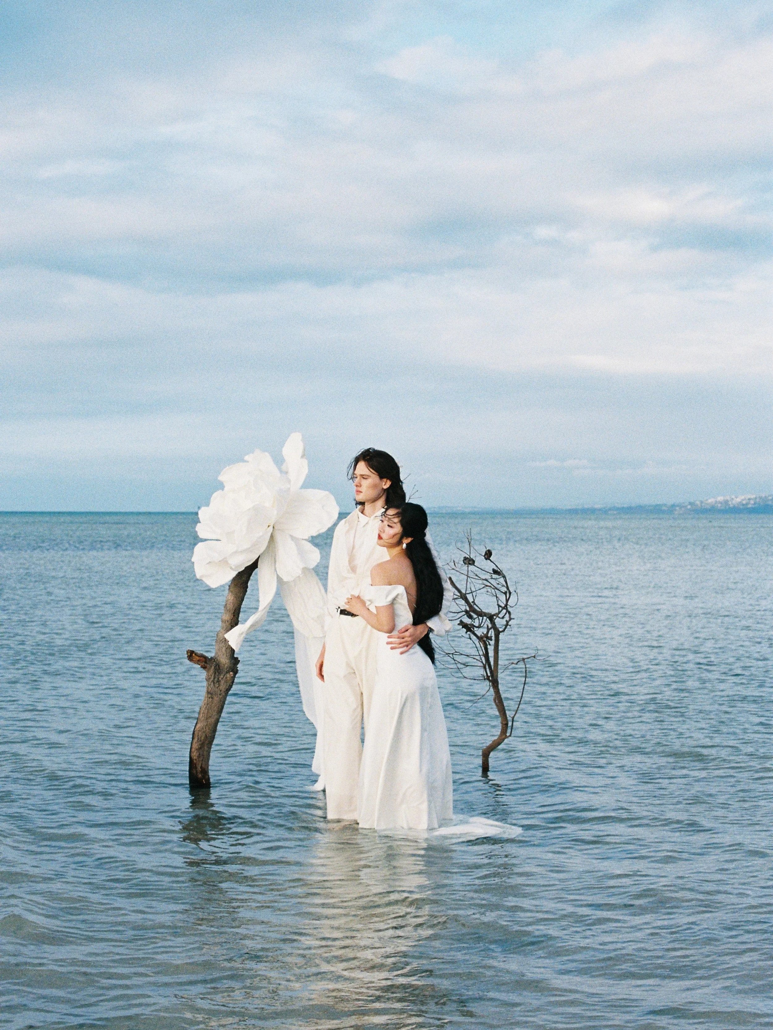 Two women standing in shallow water at the beach, surrounded by driftwood and a large white flower, under a cloudy sky.