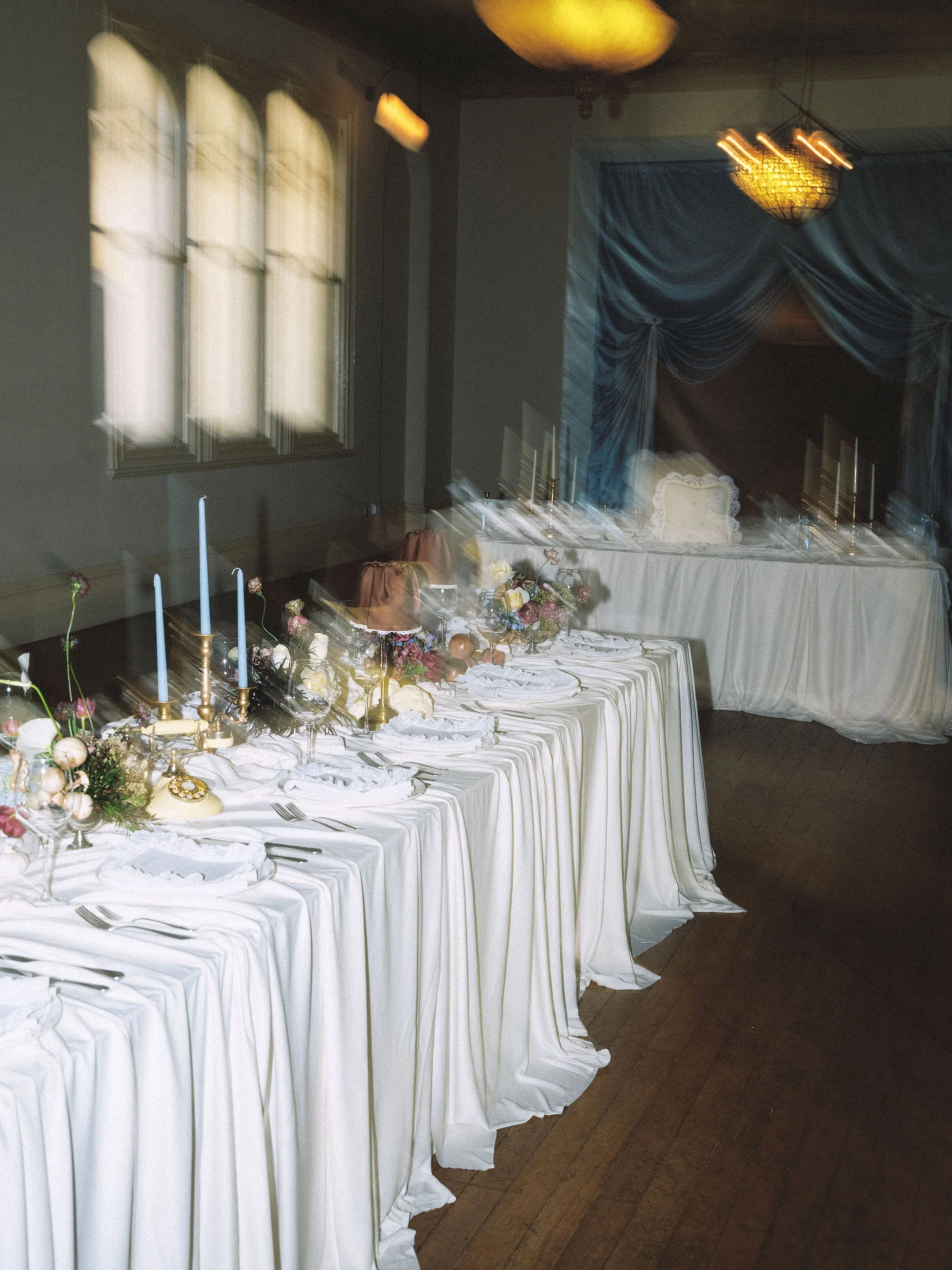 A long table with white tablecloths set for a formal event, decorated with flower arrangements, tall candles, and ornate gold candlesticks in a room with large arched windows, blue drapes, and a chandelier.