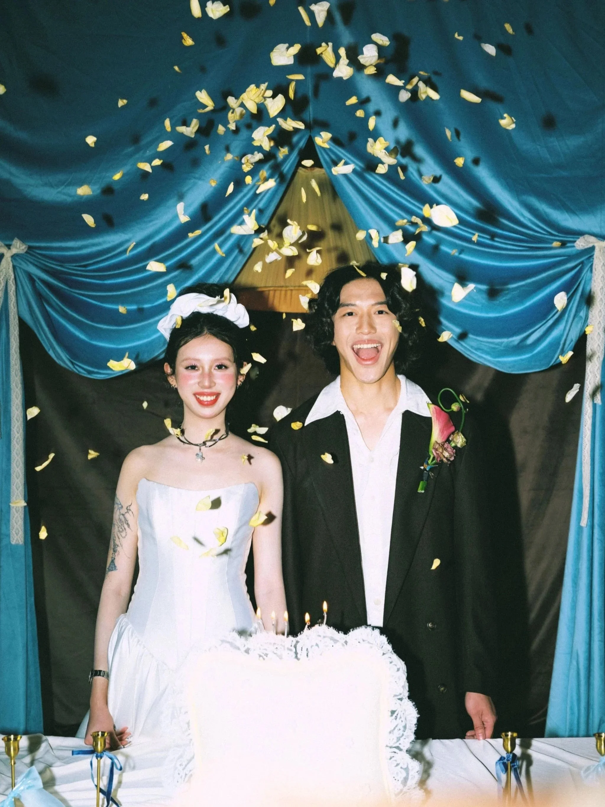 A couple dressed in wedding attire smiling under yellow confetti, standing behind a table with candles and a tiered wedding cake.