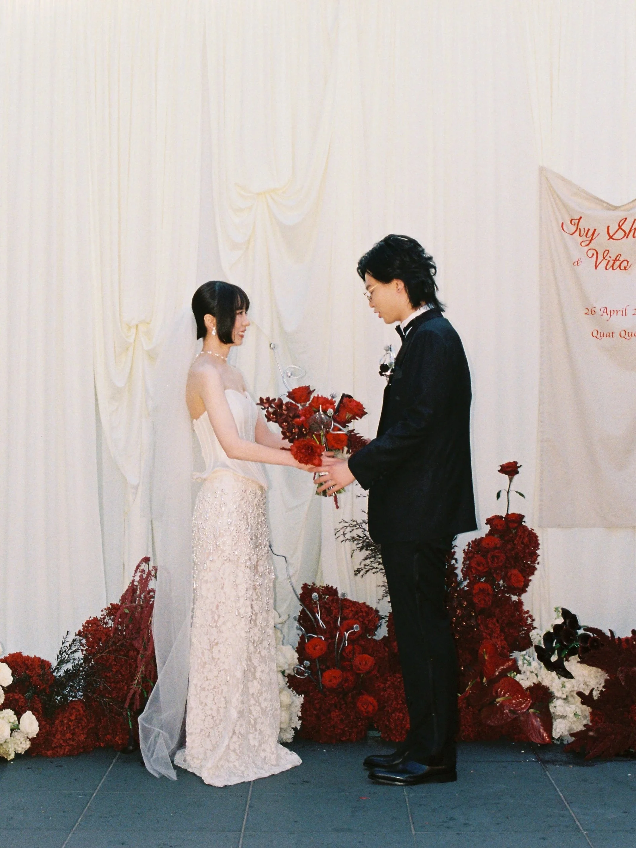 A bride and groom exchanging vows at a wedding ceremony, standing in front of a white backdrop with red floral decorations around them.