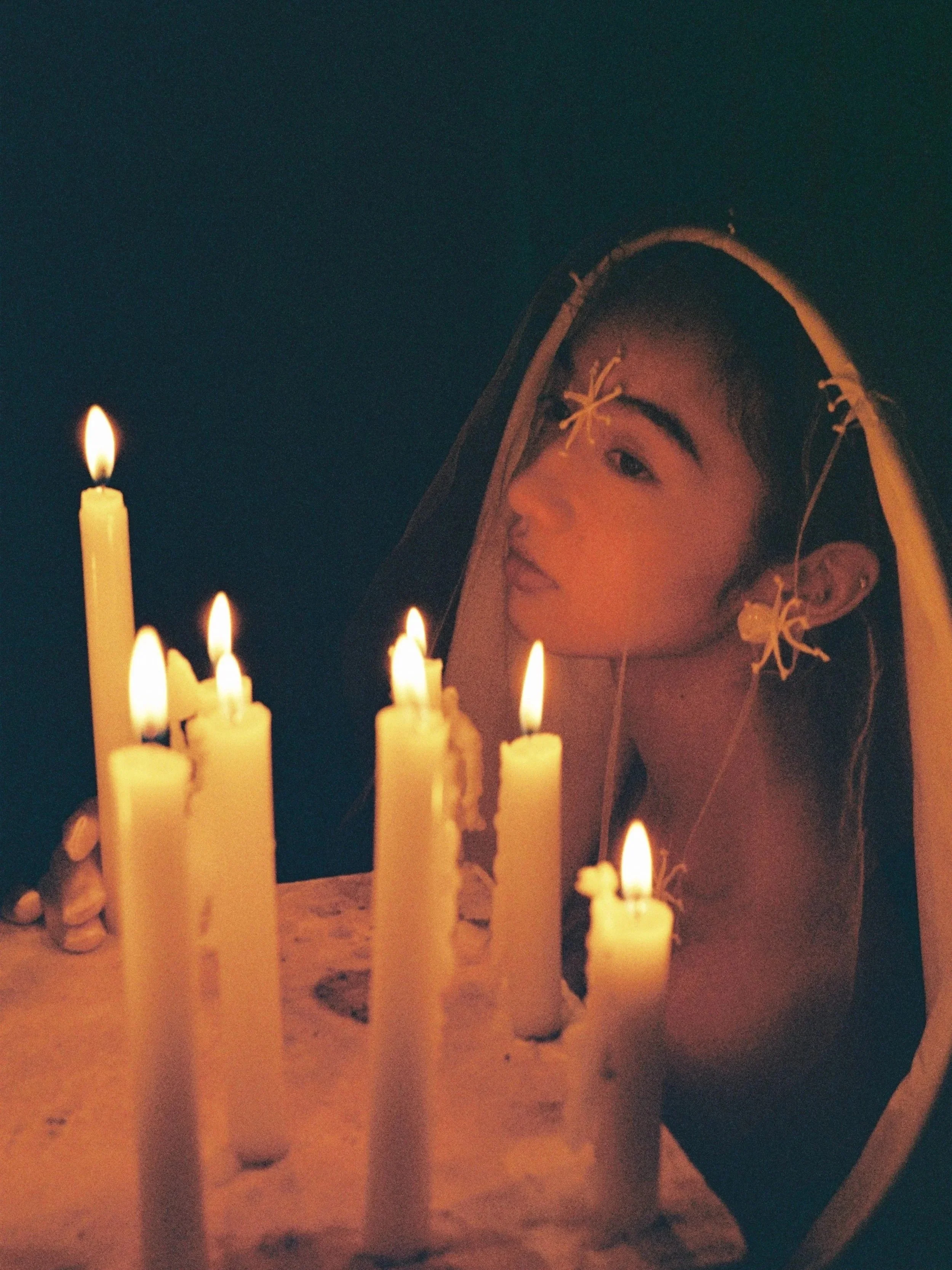 A young woman with dark hair wearing large earrings and flowers in her hair, looking at candles on a birthday cake with a dark background.