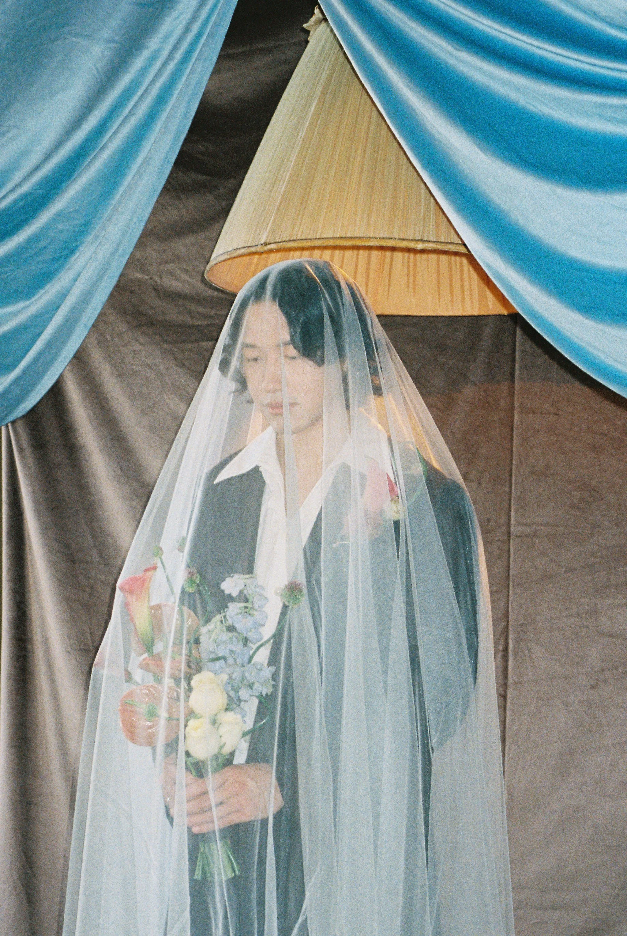 A woman with short black hair wearing a white shirt and dark jacket is behind a transparent veil, holding a bouquet of flowers.