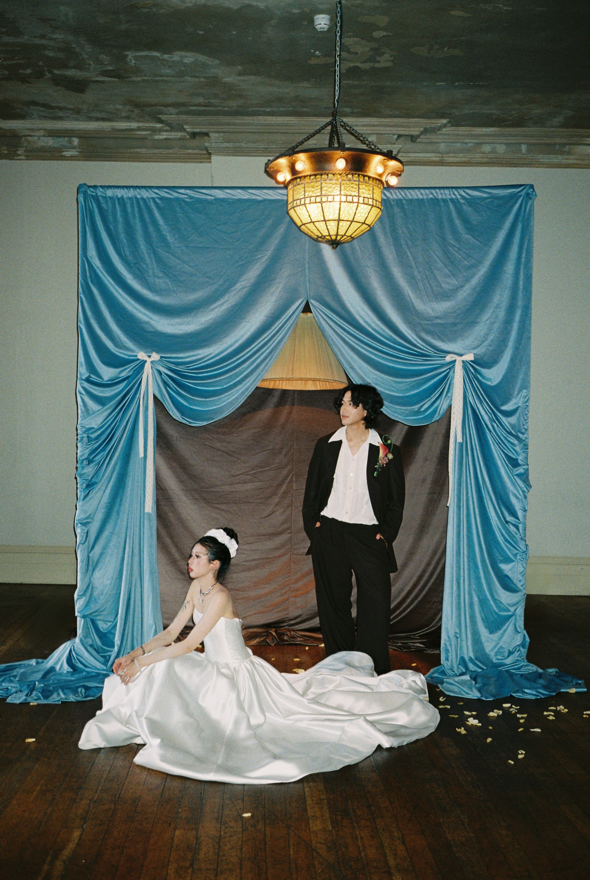 A scene with three women dressed in vintage-style clothing, set against a backdrop of blue drapes, a brown curtain, and a large tiled ceiling light. One woman is seated on the floor in a white dress, another is standing in a black suit with a flower on her lapel, and the third woman with glasses and a dark top stands behind her. The floor is wooden and scattered with white flower petals.