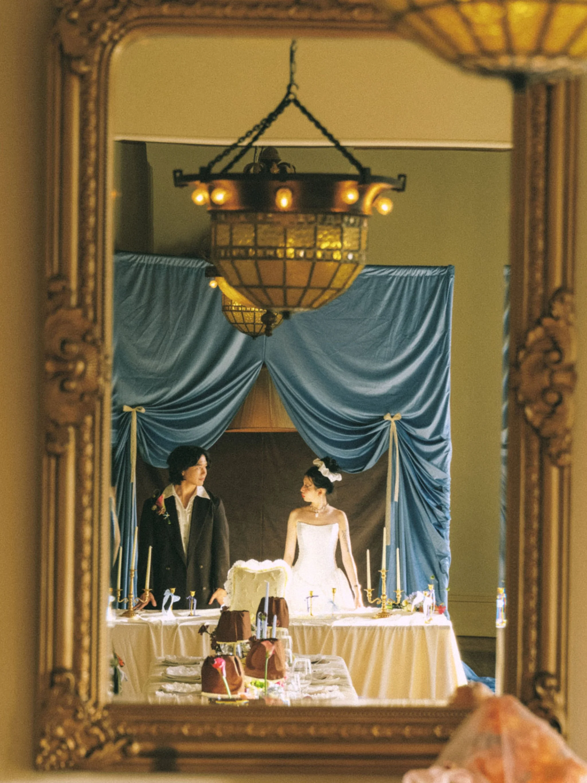 A mirror reflects a scene of a wedding reception with a bride and groom standing behind a table with a wedding cake and desserts. The scene includes blue curtains, a chandelier, and a decorated banquet table with candles and desserts.