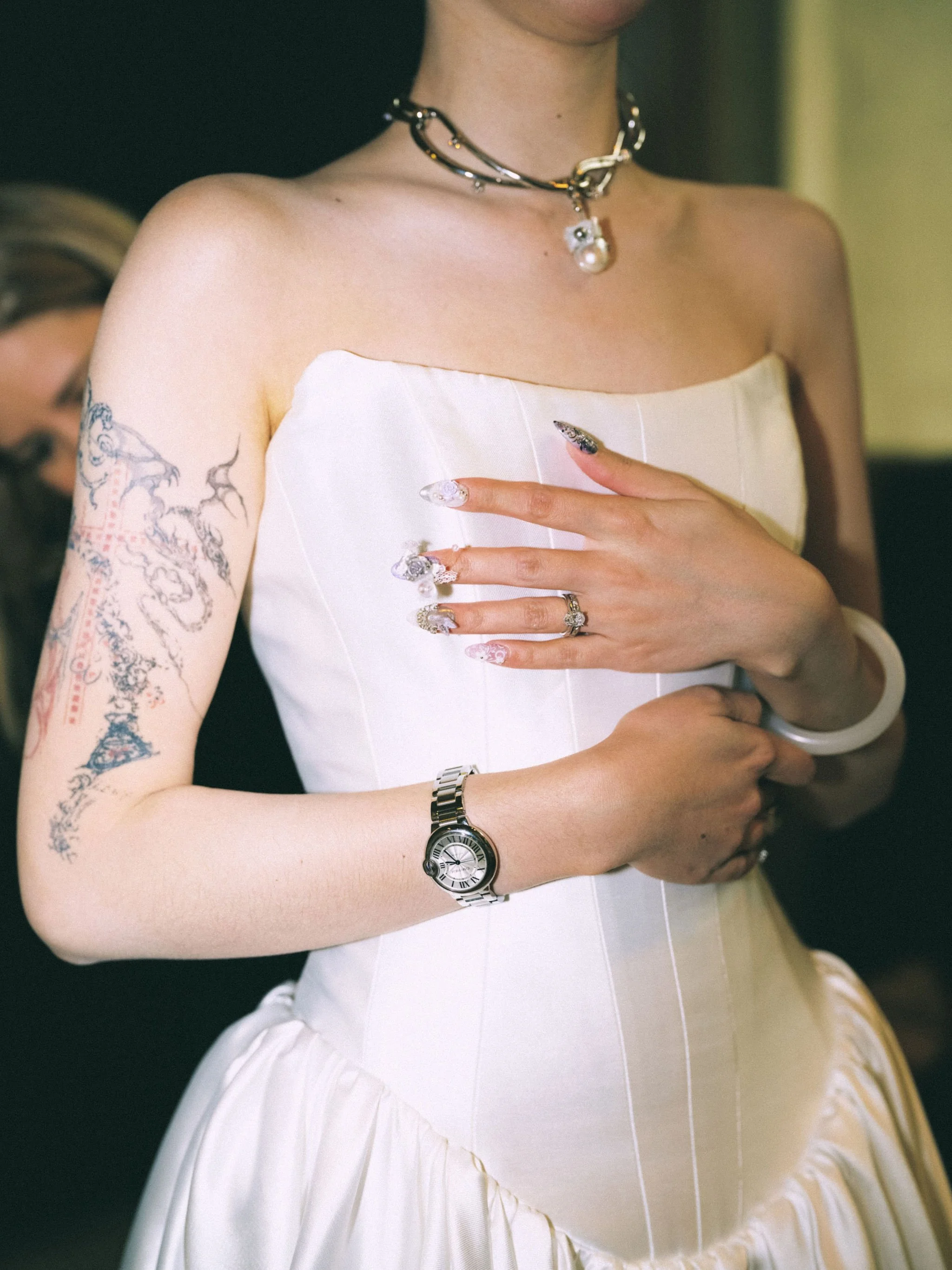 Close-up of a woman wearing a strapless white dress, showing tattoos on her upper arm, jewelry including a choker and rings, and a watch on her wrist. She is placing her hand over her chest.