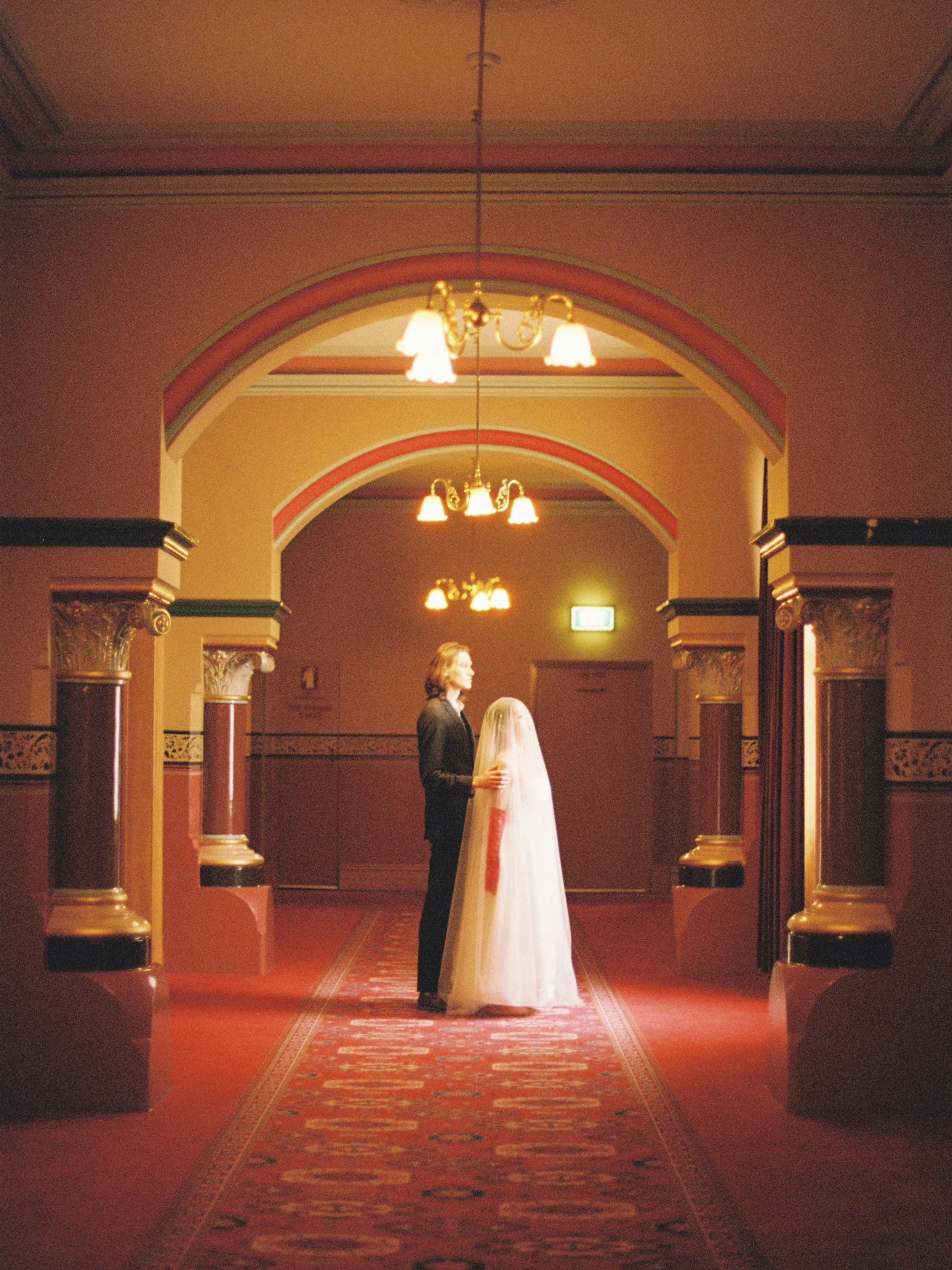 A man in a dark suit standing beside a woman in a white wedding dress in an ornate hallway with arches, chandeliers, and a decorated carpet.