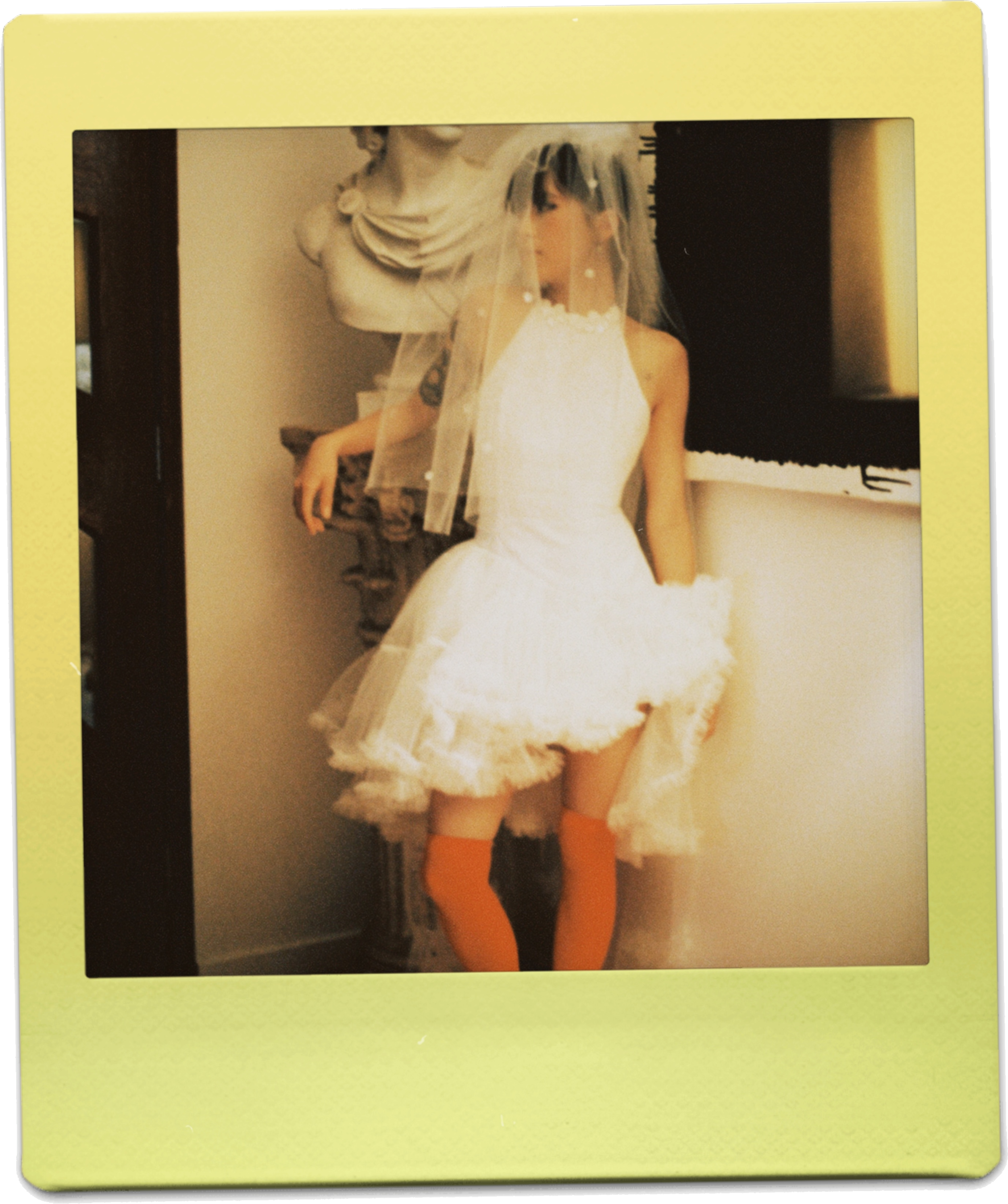A woman in a white dress with a veil and orange stockings, possibly a bride, standing indoors near a wall and a window with dark curtains. A bust sculpture is mounted on the wall behind her.