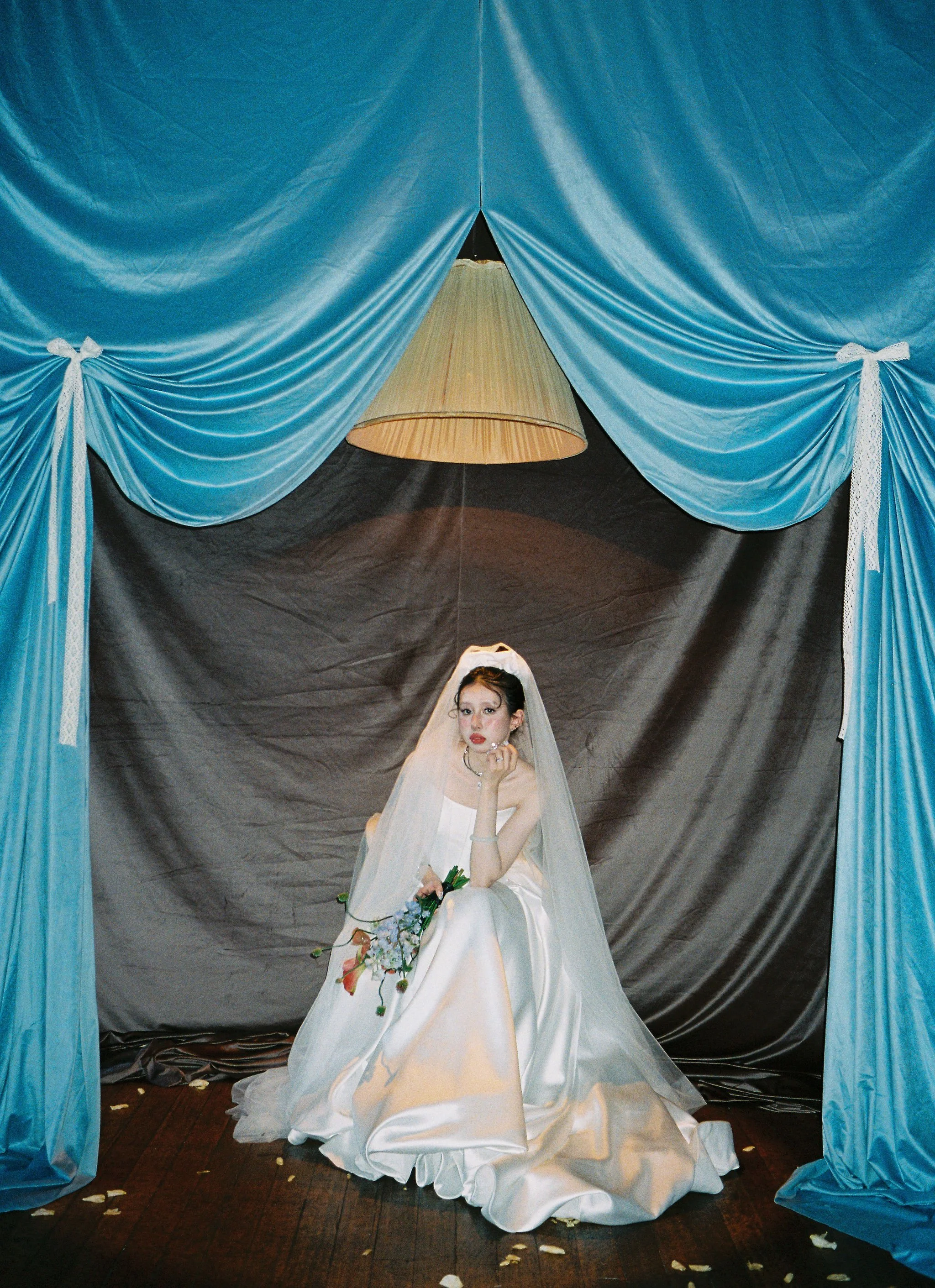 Young woman dressed in a white wedding gown sitting under blue curtains with a large beige lamp hanging above her. She has dark hair and makeup, holding a bouquet of flowers in her lap, with a thoughtful expression.